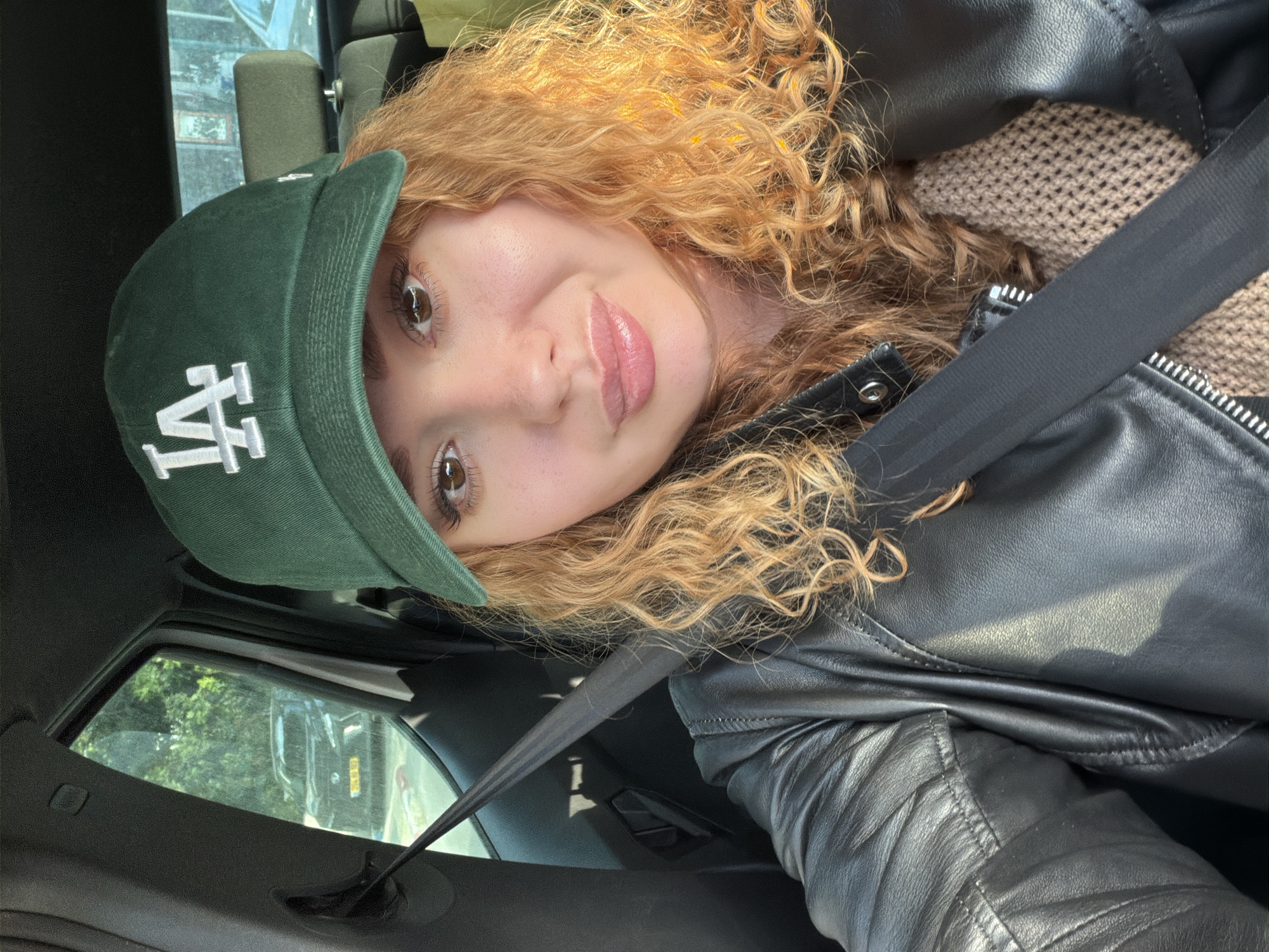 A person with light skin, curly blonde/red hair, and brown eyes is in a car, wearing a green baseball cap with a white "LA" logo and a black leather jacket over a beige knitted sweater. The person is also wearing a seatbelt. The interior of the car is visible, showing the dashboard and window. Outside the window, the environment is blurry and indistinct, but appears to be a city street with buildings and cars. It is impossible to determine the location in the city of Tel-Aviv, Israel.