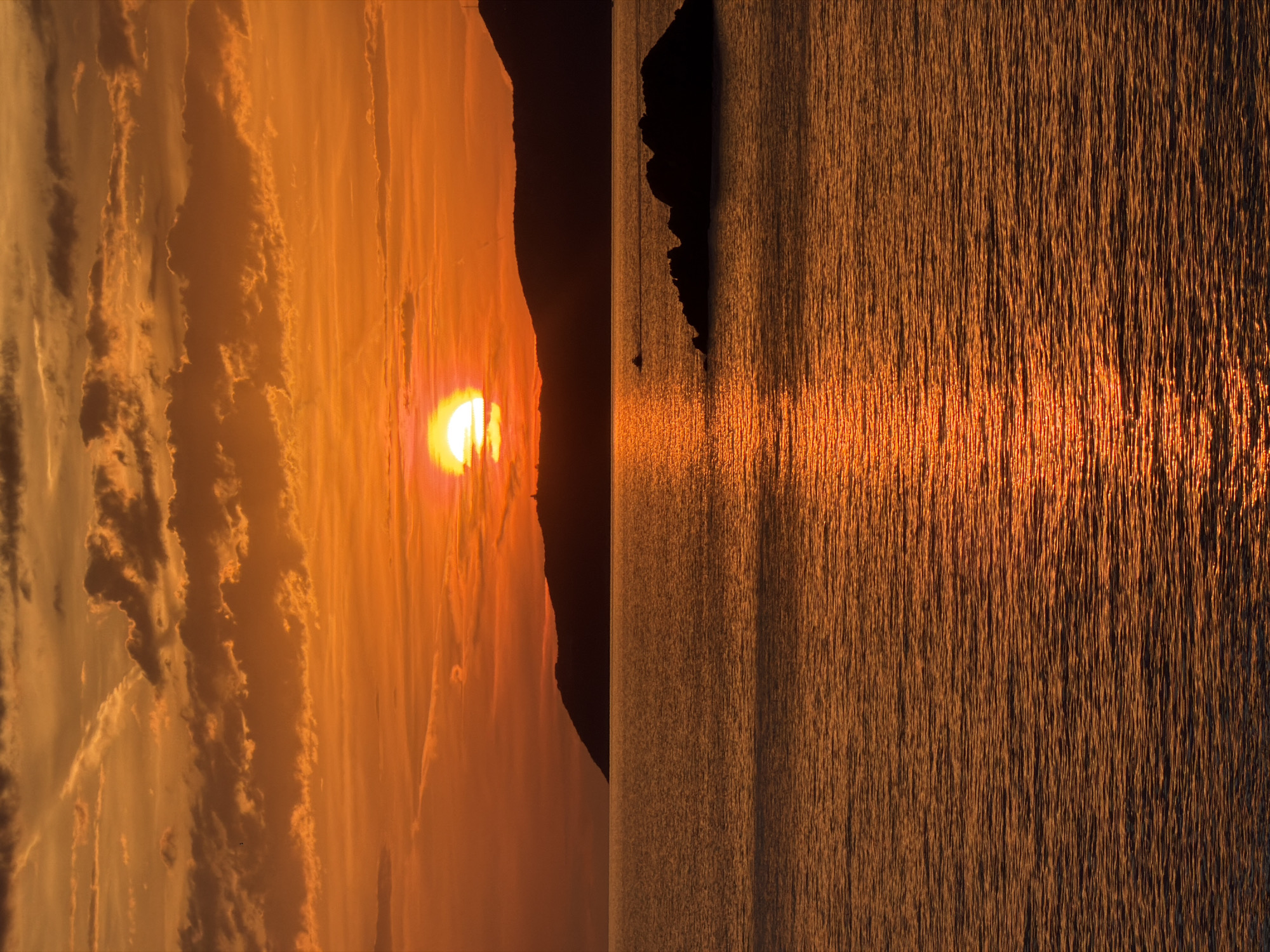 This image depicts a sunset over the Adriatic Sea at Perazića Do, Montenegro. The sky is saturated with intense orange and reddish-orange tones, featuring scattered clouds of varying density across the upper portion. The sun, appearing as a brilliant yellow-orange semi-circle, is partially obscured by the silhouette of a dark, distant hill on the horizon. Another silhouetted hill rises to the right, with a faint, slender structure visible at its peak. The expansive water surface below reflects the vibrant sky, creating a shimmering path of broken light extending from the horizon towards the foreground. Small ripples are visible across the water's surface. In the mid-ground, a dark, rocky islet is positioned towards the right, and a small, dark silhouette of a boat or watercraft is visible further to the left. No individuals are present in the scene.