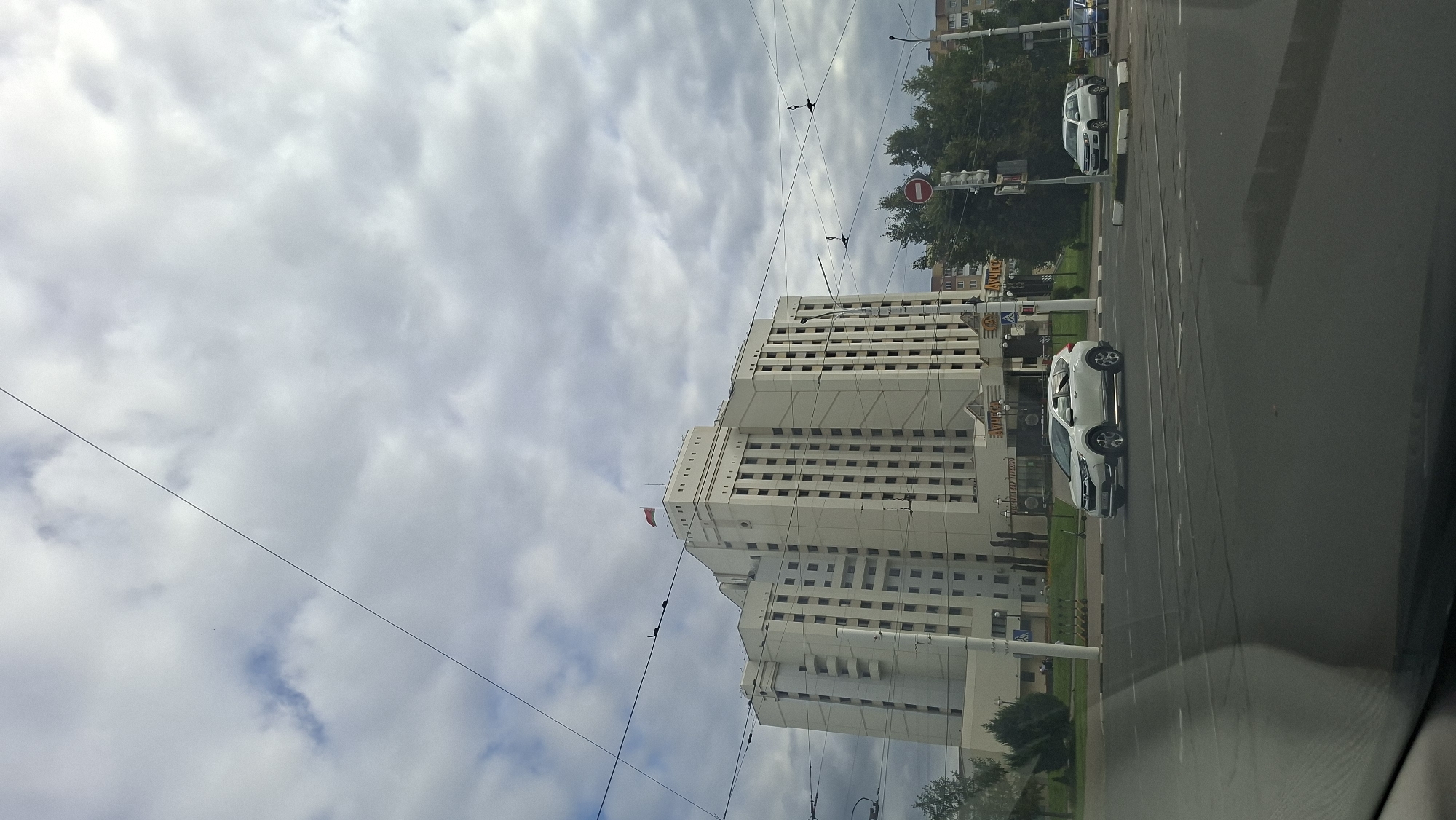A white, multi-story building with a red and green flag on top dominates the scene. The building is rectangular with many windows and a slightly stepped profile at the top. Power lines crisscross the sky. In front of the building, a white SUV is parked on the street. Another white SUV is visible in the background. A traffic light with a red circle sign is also present. The scene appears to be a street in a city with some trees and other buildings visible in the distance. The sky is overcast with clouds. The location is the Vitebsk City Executive Committee building in Vitebsk, Belarus.