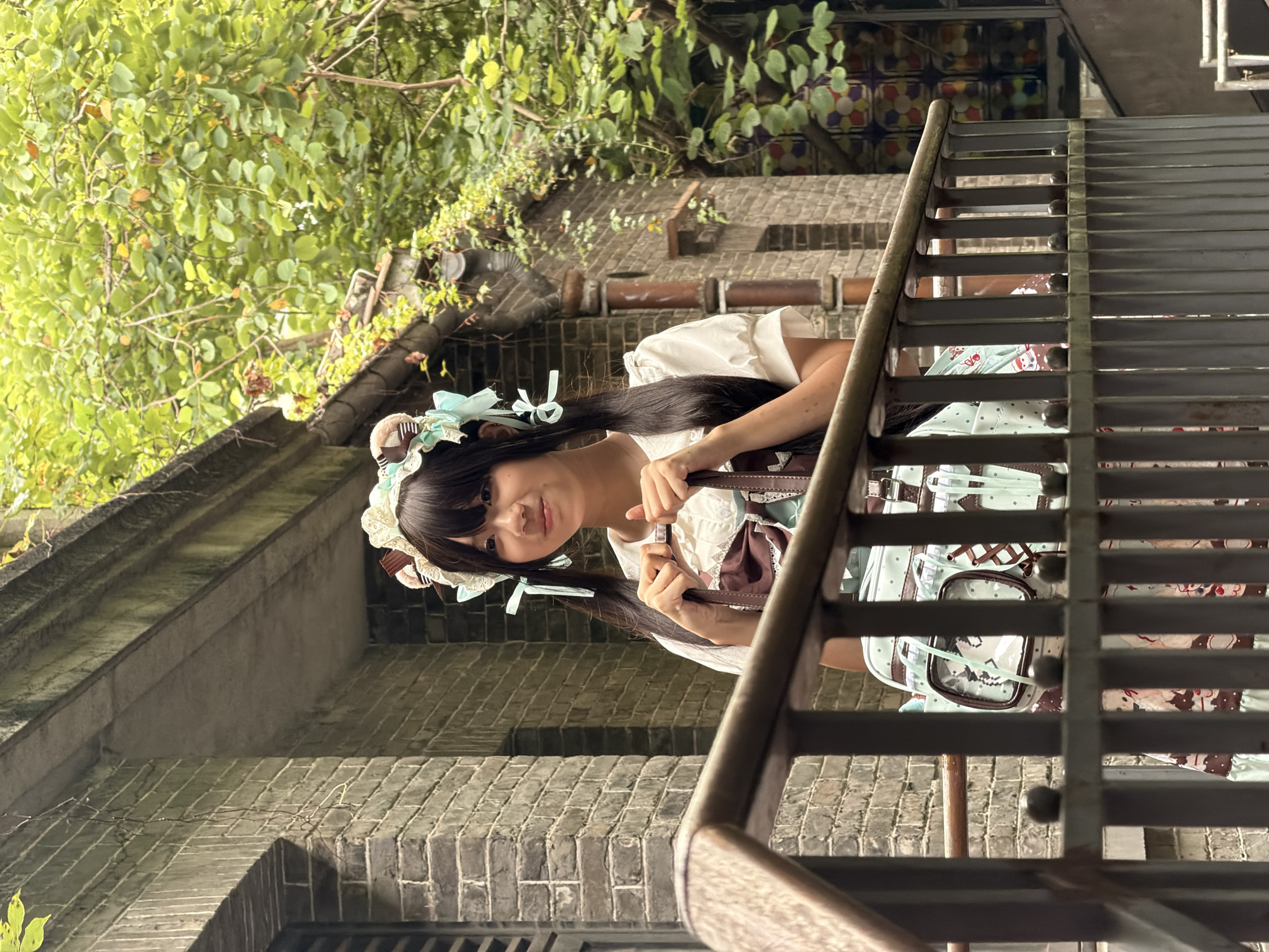 An Asian female, appearing to be in her late teens or early twenties, stands behind a metal railing. She has black hair styled in pigtails and wears a white dress with puffed sleeves, a brown and white pinafore with a large bow, and a decorative headband with pastel-colored bows and embellishments. She is holding a brown handbag. Her lower body is partially obscured by the railing and her patterned skirt, which features whimsical designs. The background consists of a brick building with ivy growing on it, and lush green foliage. In the background, there are also visible jars filled with colorful objects. The overall scene suggests an outdoor setting, potentially a garden or courtyard area, within a city environment. The architectural style of the building, characterized by exposed brickwork and decorative eaves, could be indicative of older districts or traditional structures. While the exact location within Chancheng District, China cannot be definitively determined from the image alone, the presence of such architectural elements and vegetation aligns with the diverse urban landscapes found in many Chinese cities.