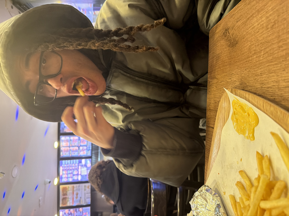 A person with dreadlocks, glasses, and a hooded jacket eats a French fry. They are at a wooden table, with a tray holding the remaining fries and a dollop of cheese. The background features a menu board illuminated with blue lights. A second person is partially visible in the background, seated and wearing dark clothing. The photo appears to be taken indoors, possibly at a restaurant.