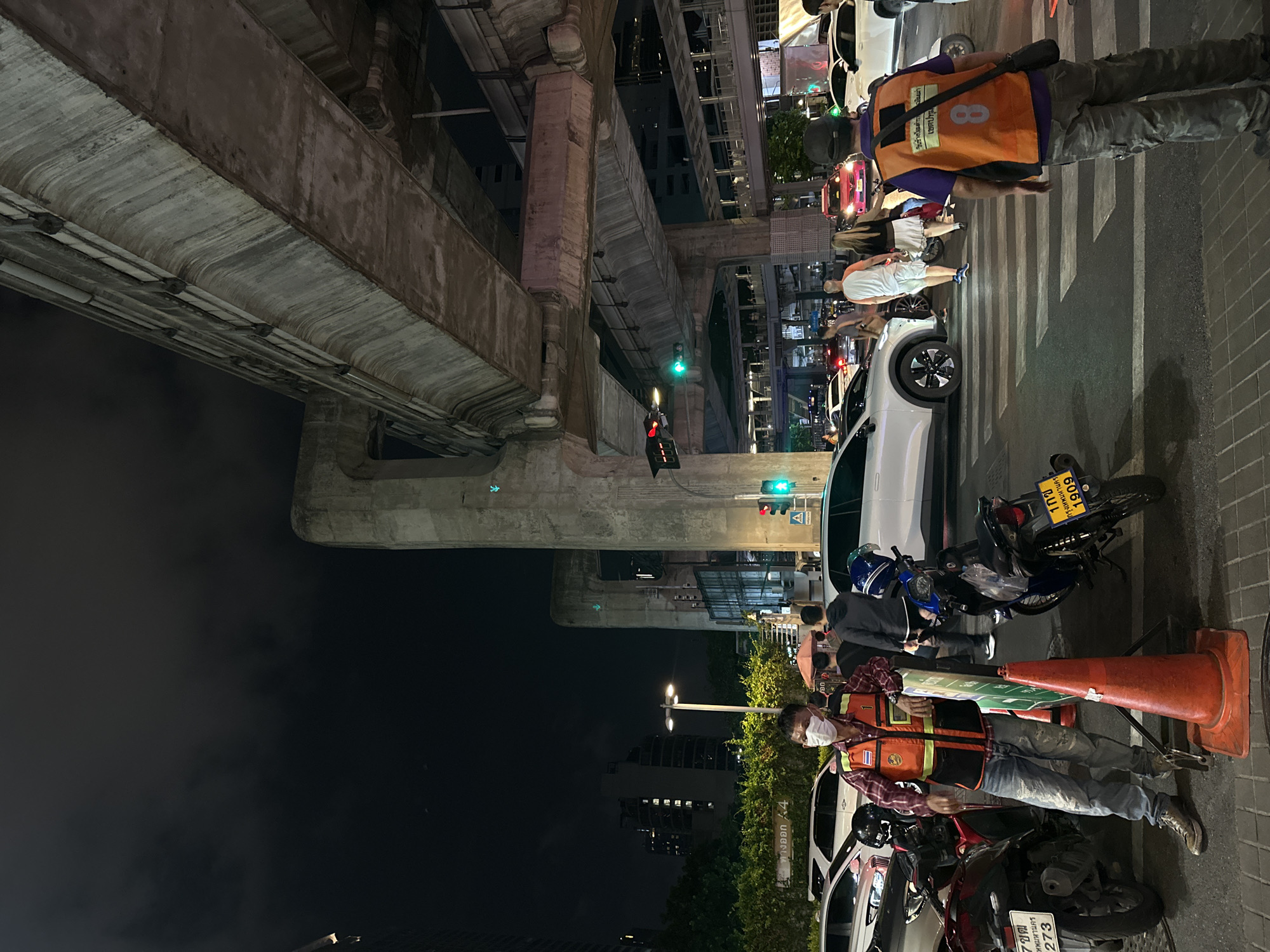 The image captures a nighttime scene in Bangkok, Thailand, beneath an elevated train track structure.
People and Activities: Pedestrians are crossing the street at a crosswalk. Some are walking, and others are waiting, with a traffic controller present. Another person in a safety vest is present near parked motorcycles.
Subjects and Objects: A white car is stopped at the crosswalk, along with a motorcycle. Orange traffic cones are also visible. The backdrop includes a building and streetlights. One motorcycle has the Thai license plate "1 กช 6061 กรุงเทพมหานคร", and another has "1กฮ 273". There is a sign that says "ทางออก 4".
Environment: The setting is an urban environment with elevated train tracks overhead. It is dark, but the area is illuminated by streetlights and traffic signals. The photograph is taken at ground level, looking upwards.