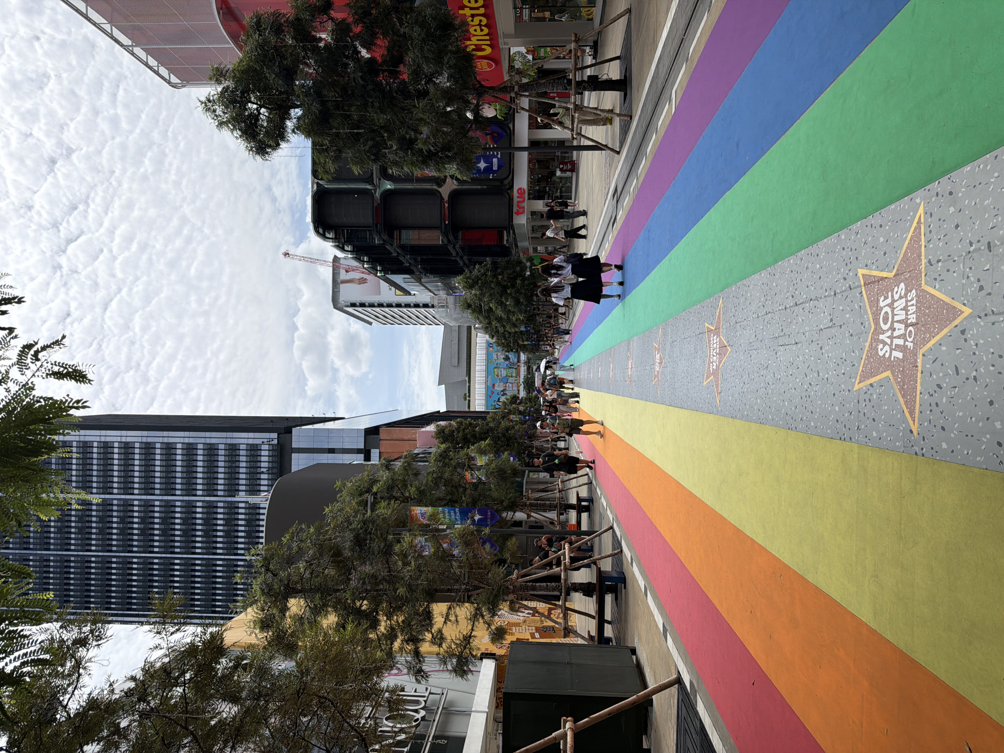 The image shows a street in Bangkok, Thailand, decorated with a rainbow-colored path. The path, which appears to be a pedestrian walkway, features stripes of red, orange, yellow, green, blue, and purple. Several people are walking on the path, some dressed casually and others in school uniforms. Buildings line both sides of the street, with commercial establishments and tall skyscrapers visible. Trees are planted along the walkway, with some trees pruned and supported by wooden stakes. In the background, a construction crane is visible, suggesting ongoing development in the area. The sky is partly cloudy with a textured cloud pattern. Some stars with text are embedded in the grey parts of the pathway, with one visible star reading "STAR OF SMALL JOYS". The overall impression is of a vibrant and modern urban environment with a festive decoration.