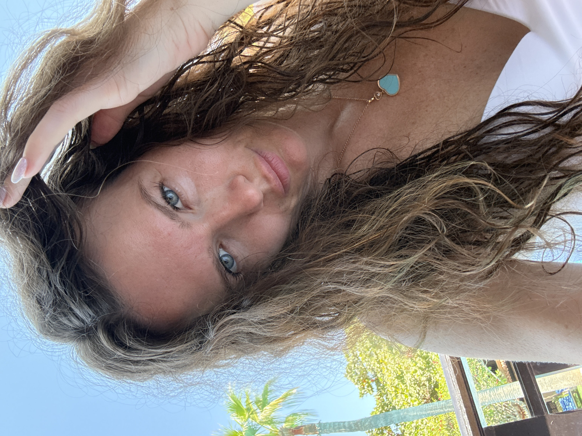 A young woman with blue eyes and long, wavy brown hair is taking a selfie outdoors. She is wearing a white top and a necklace with a heart-shaped turquoise pendant. Her left hand is raised, touching her hair. The background shows a clear blue sky, palm trees, and greenery, suggesting a tropical or subtropical location. The lighting indicates it is daytime. Based on the flora, it is likely the woman is in Dubai, United Arab Emirates.