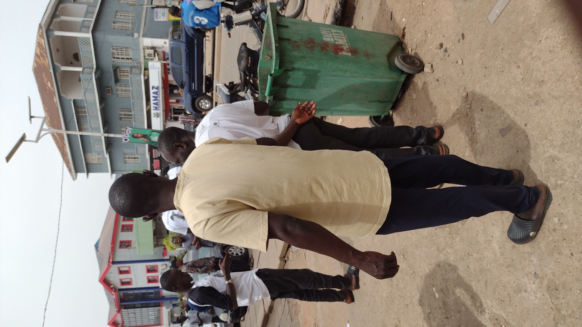 The image, rotated 90 degrees clockwise, captures a bustling street scene in Jos, Nigeria, likely during daytime under an overcast or hazy sky. The foreground is dominated by several men, one prominently in a yellow short-sleeved shirt, facing away from the viewer. To his right, another man in a white short-sleeved shirt leans or sits beside a large, green wheeled garbage bin. This bin has "HAPPY MILLY" written in white capital letters on its side, along with some red markings.
In the mid-ground and background, more people are visible, some standing and others possibly engaged in commercial activities. Vehicles are present, including a dark blue pickup truck parked near a building, and a motorcycle or scooter next to the garbage bin.
The setting features multi-story buildings lining the street. One prominent building on the left has several balconies and a distinctive rusty-colored roof, while another building in the background displays a red and white facade. Street infrastructure, such as a street light pole, is also visible. The ground appears to be a mix of dirt and paved areas.
Visible text includes a sign reading "HAMAZ" affixed to a building, and a vertical banner with "Opay" displayed on another structure. A person near the blue pickup truck is wearing a blue jersey or shirt with the number "3" on its back. The overall impression is one of everyday life in an active urban environment.