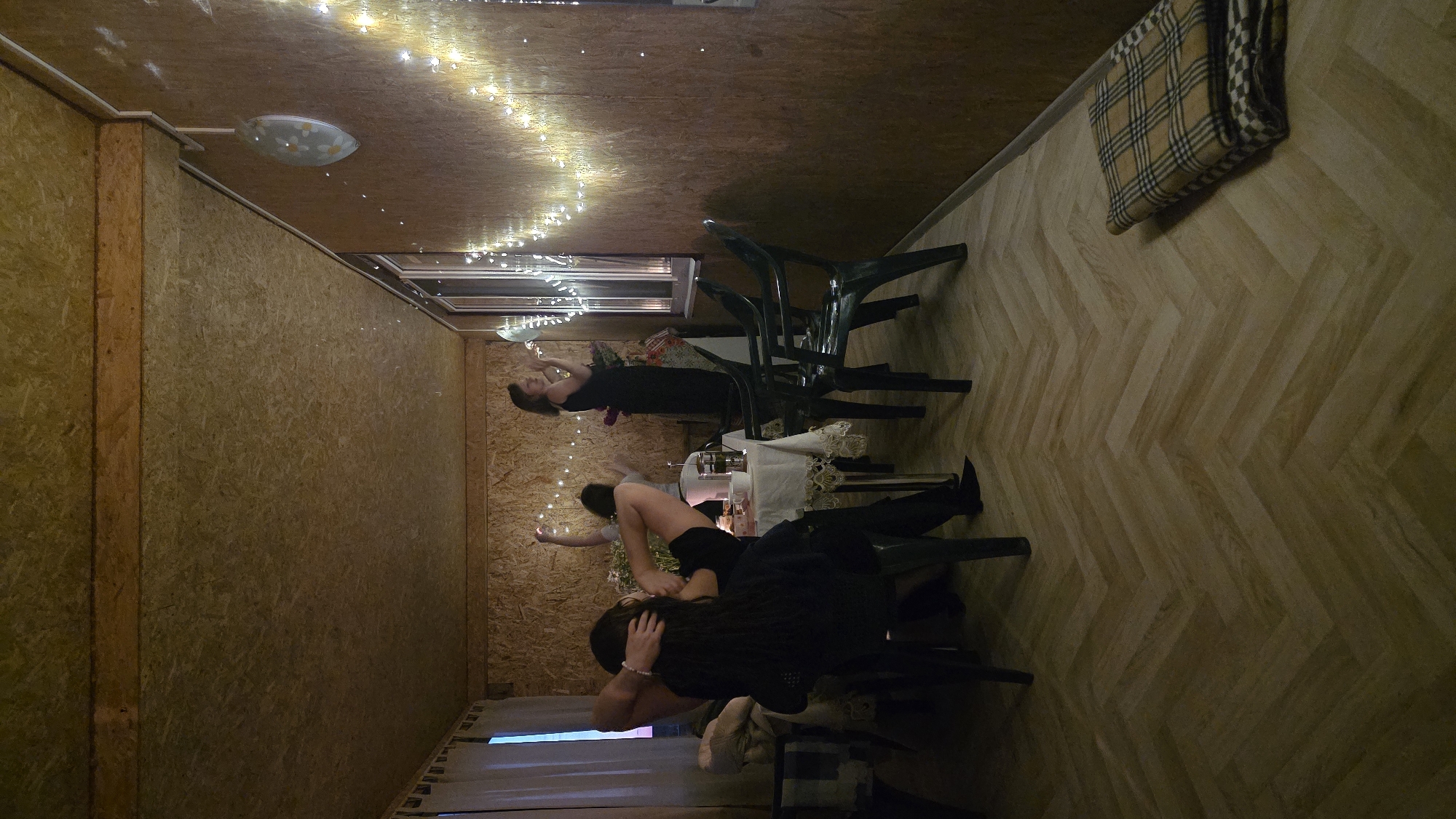 The image depicts an indoor scene, likely a room or enclosed porch with wooden walls and ceiling. The room is lit by string lights. In the foreground, a person is seated, facing away from the camera, with their legs propped up. To their left, another person is visible, also seated. In the center of the room, a table is set with a white tablecloth, teacups, and a teapot. Three other individuals are standing or moving around the table. One person is standing behind the table, another is near the window and appears to be holding something, and a third is in the background with their arms raised. The floor is covered with a herringbone pattern wood laminate. A folded blanket with a plaid pattern is on the floor in the right foreground.