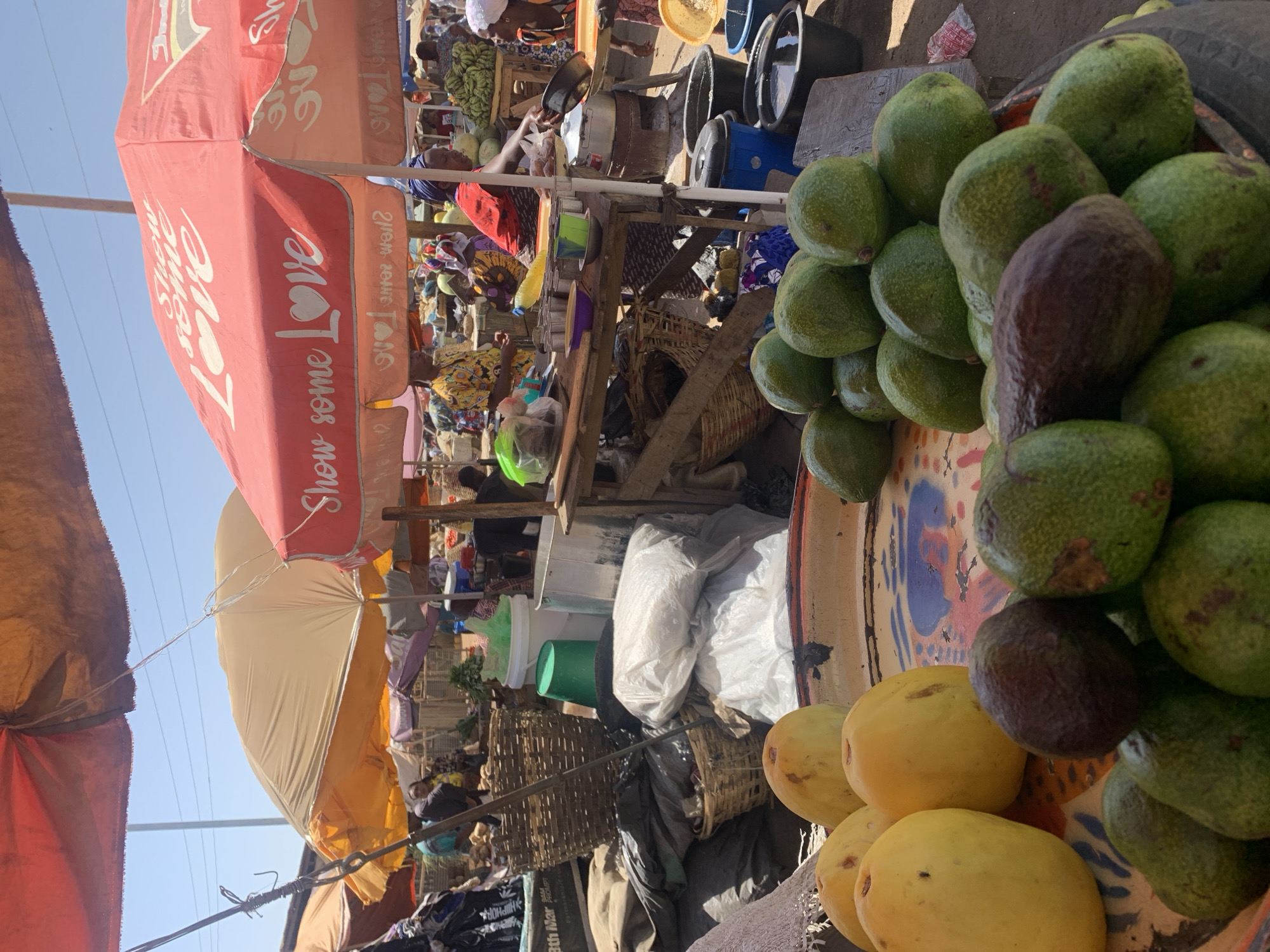The image depicts a bustling outdoor market scene in Rafin Par, Nigeria. In the foreground, a tray overflowing with green avocados and yellow fruits (possibly mangoes) is prominently displayed. Further back, vendors and customers are visible amidst stalls cluttered with various goods. The scene is partially shaded by colorful market umbrellas with "Show some Love" printed on one. Clothing, baskets, plastic containers, and bags are scattered throughout the stalls. The overall environment appears crowded and active, typical of a busy open-air market.