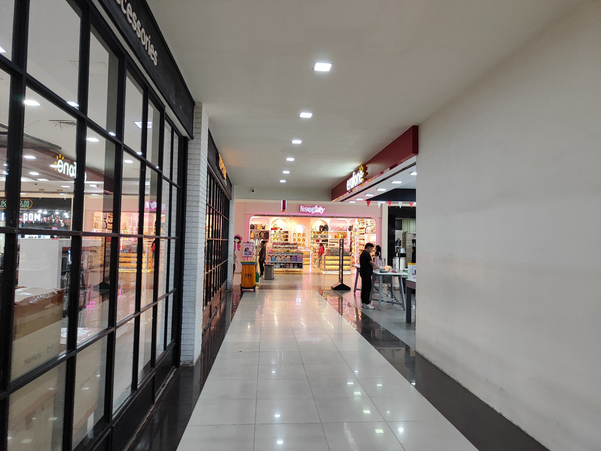 The image depicts a well-lit interior hallway of a shopping mall. The hallway features polished floors with a reflective surface that mirrors the ceiling lights and the shops lining the corridor. On the left, large windows with black frames reveal the interior of a shop displaying various items, including boxed goods. The shop's signage includes the word "Accessories" in white lettering against a dark background. Further down the hallway, other shops are visible, including one with pink and white signage that reads "Naughty." A few individuals are present in the hallway. One person, wearing a black shirt and pants, stands near a table with items displayed. Another person, in a striped shirt, is seen walking away. In the distance, a person in a pink shirt can be observed. The architecture suggests a modern retail environment with ample lighting and clean lines. The scene appears to be located in Sempusari, Indonesia, as indicated by a textual reference.