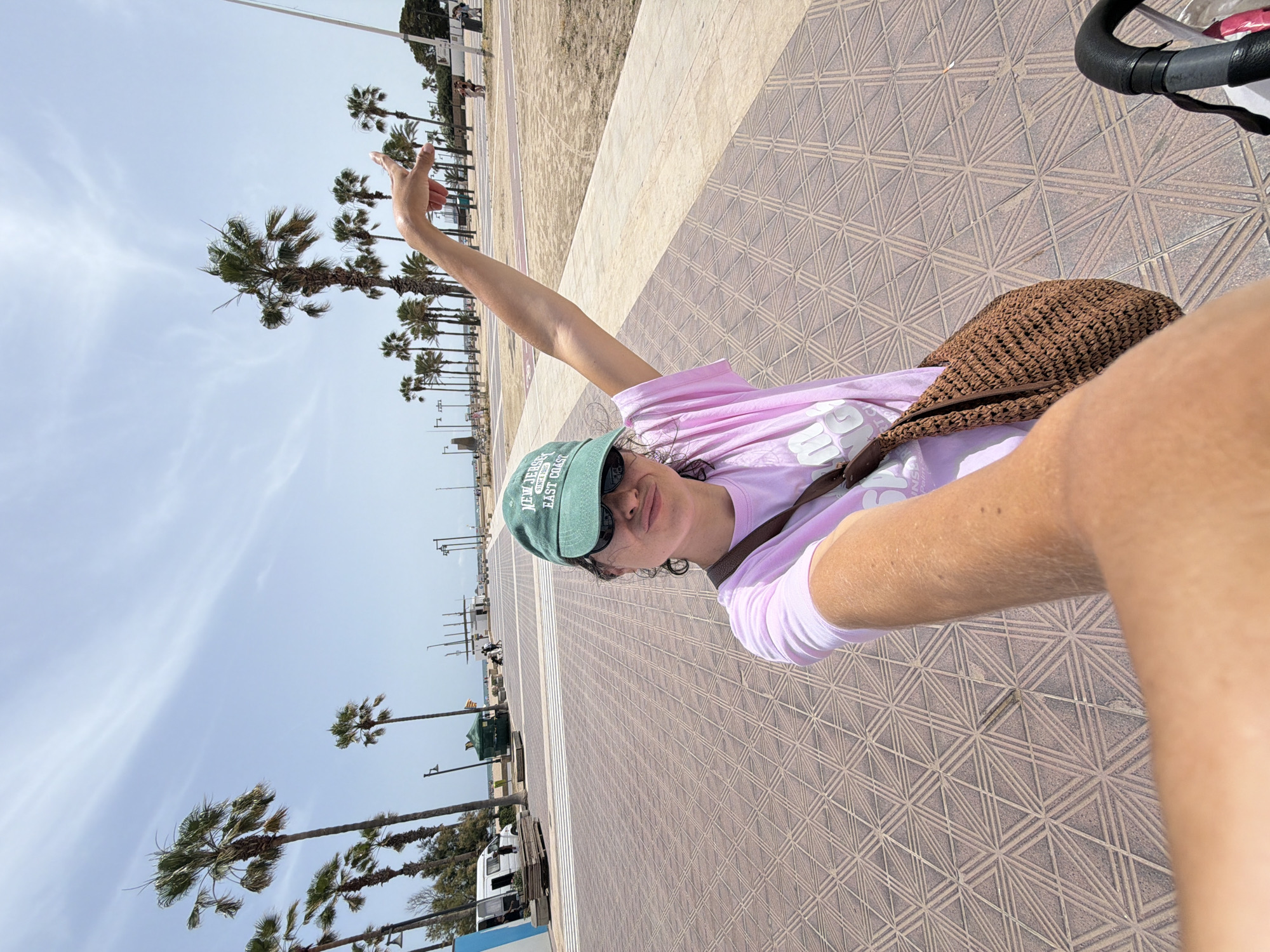 In an outdoor, daytime scene, a young woman with curly dark hair, wearing a pink t-shirt, sunglasses, and a green baseball cap, stands on a patterned tiled walkway. She is taking a selfie and playfully making a "duck face." Her right arm is raised, and she points her index finger upwards towards the sky. A brown woven bag hangs from her left shoulder. The background features a row of palm trees, a sandy area, a light blue sky with some wispy clouds, and buildings and streetlights in the distance. The overall impression is a casual, sunny day outing in a coastal or tropical location.