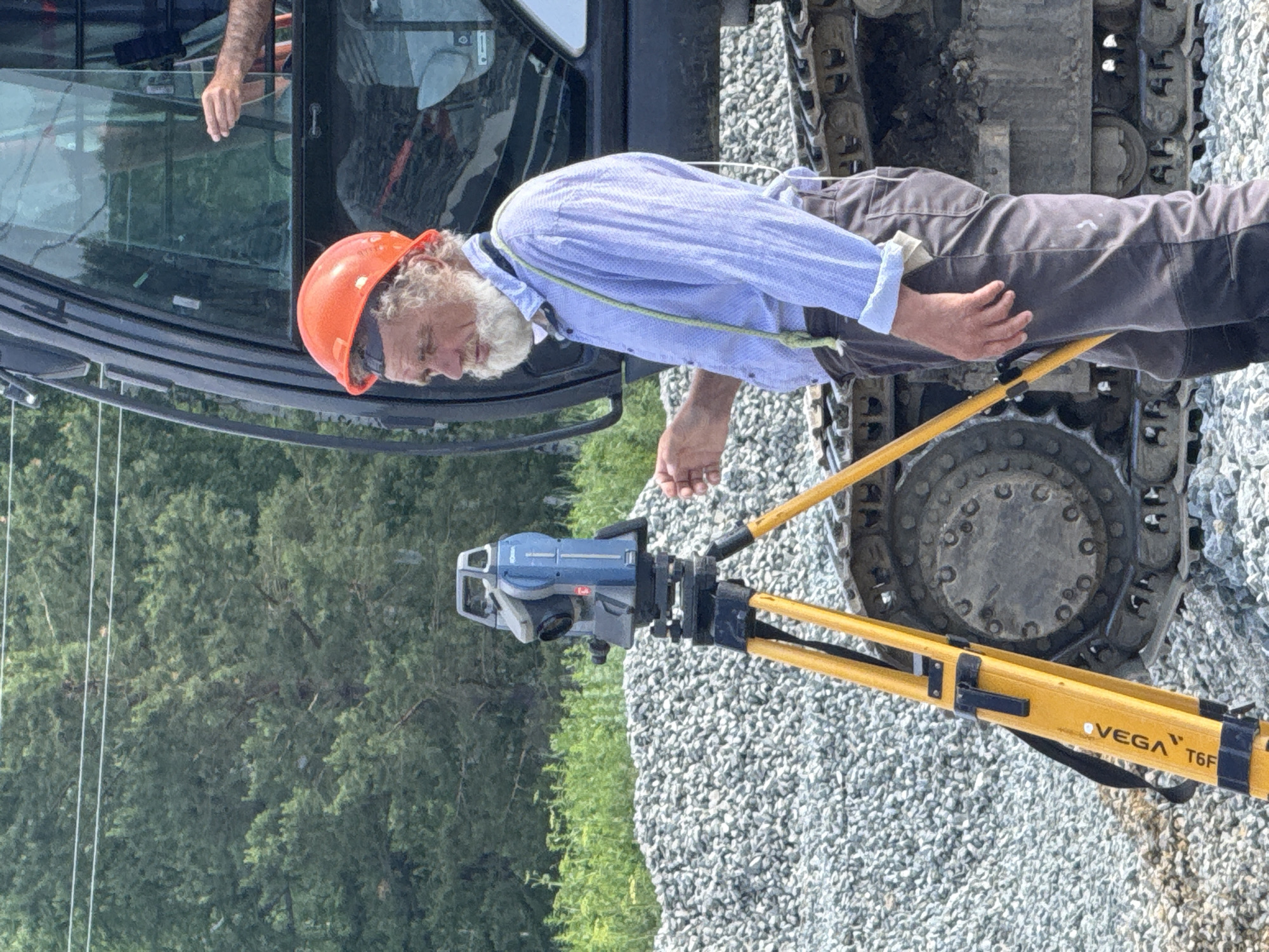 An older man with a white beard and a red hard hat stands near a large construction vehicle. He is wearing a light blue collared shirt and dark pants, with green suspenders. In front of him is a blue and grey surveying instrument mounted on a yellow tripod. The man is holding a hand up and appears to be interacting with the instrument or giving instructions. The ground is covered in grey gravel. In the background, there is a dense forest of green trees and a power line. The scene appears to be a construction site in Leninsk, Russia.