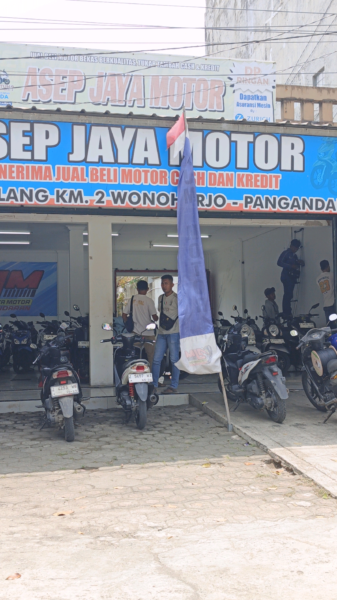 The image shows a storefront with the name "ASEP JAYA MOTOR" displayed prominently on a blue banner. The storefront appears to be a motorcycle dealership in Desa Wonoharjo, Indonesia, as indicated by the text "LANG-KM. 2 WONOHARJO - PANGANDA". Several motorcycles are parked in front of and inside the establishment. Two men are standing near the entrance, one of whom is looking towards the camera. Another man is inside the dealership, possibly a customer or employee. A large blue flag is prominently displayed in the foreground, obscuring a portion of the storefront. The area is paved with concrete tiles and there are some overhead wires visible. The overall impression is of a commercial establishment dealing in motorcycles.