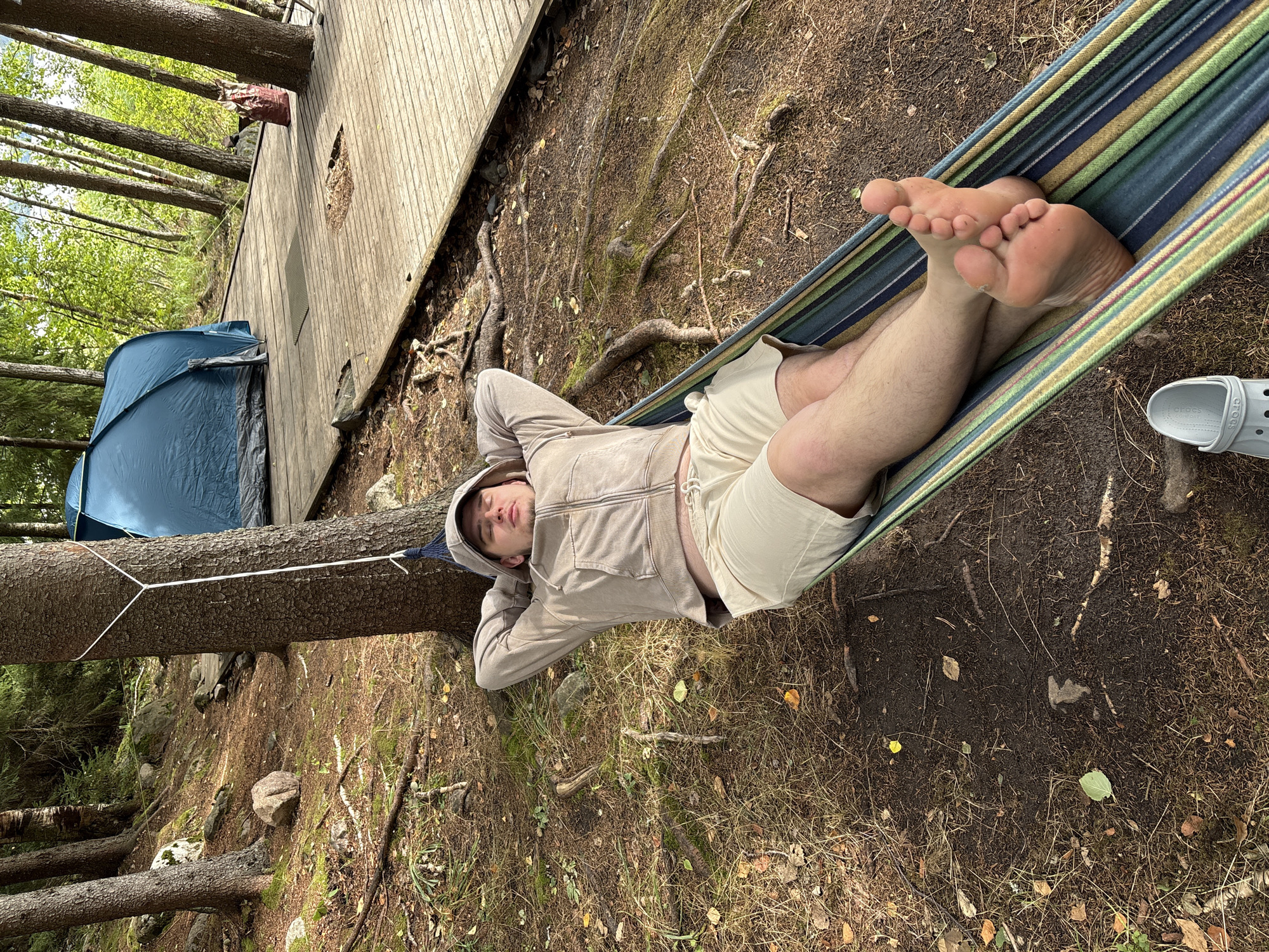 A young man reclines in a colorful hammock strung between trees in a wooded area. He is wearing a light brown hooded jacket and light-colored shorts. His eyes are closed, and he appears relaxed. The hammock is striped with green, blue, and yellow. To the left of the hammock, a blue tent is visible, partially obscured by a fallen tree trunk. Behind the tent, a wooden deck structure can be seen. The ground is covered in dirt, pine needles, and fallen leaves, with exposed tree roots and scattered rocks. Sunlight filters through the trees, suggesting it is daytime. The scene evokes a sense of peaceful outdoor recreation, likely camping or lounging in nature. There is no visible text in the image. The location context provided is Rautalakhti, Russia.