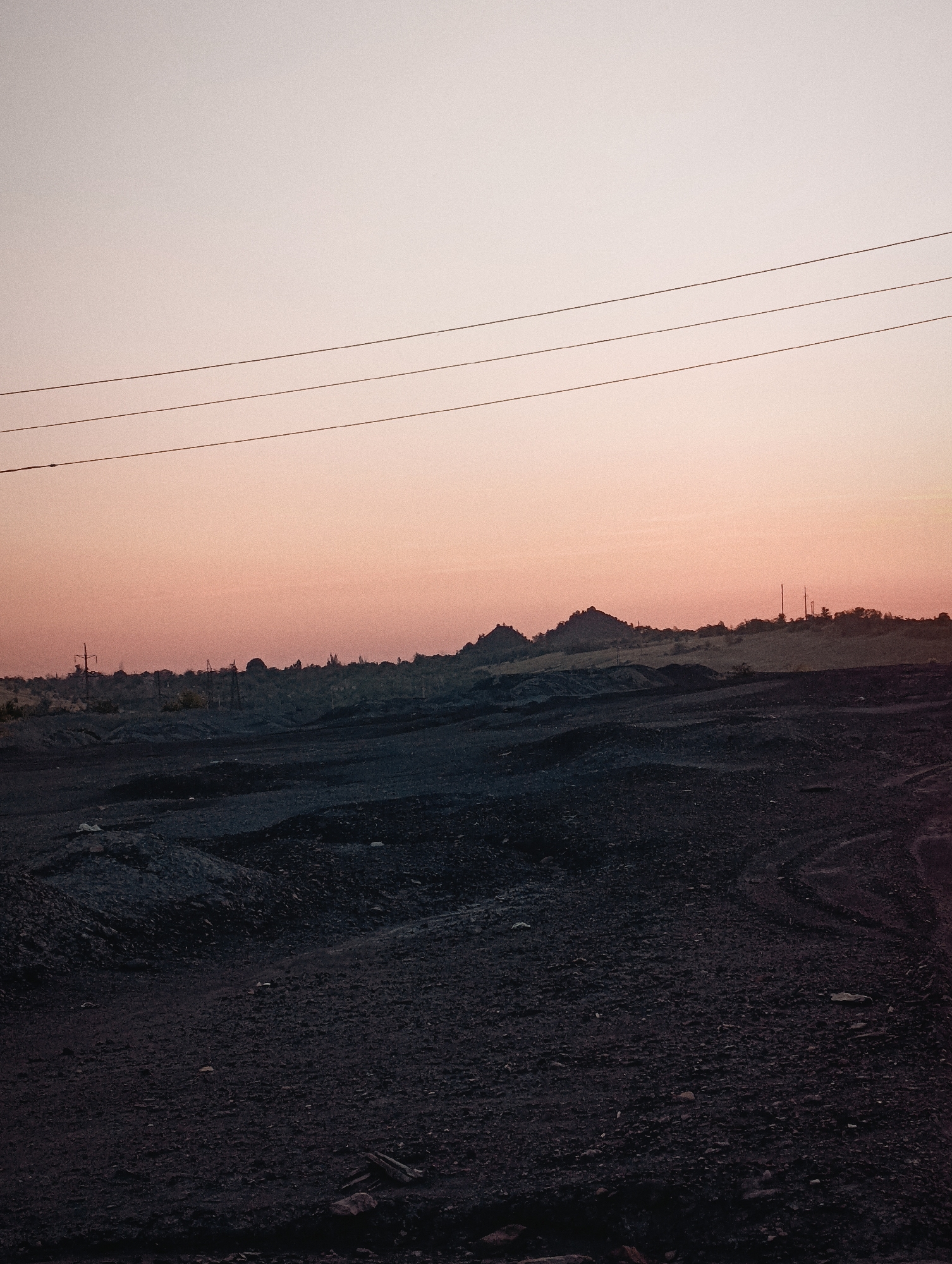 The image depicts a desolate, rocky landscape under a hazy, pink-orange sky, suggestive of either dawn or dusk. Three power lines are strung horizontally across the upper portion of the frame. The foreground is dominated by dark, rough terrain, appearing to be composed of gravel and debris, with some scattered rocks and possibly remnants of construction materials. In the middle ground, the terrain rises into undulating hills and mounds, also covered in dark material. A silhouette of a distant, jagged mountain range or cluster of hills is visible on the horizon, with a few indistinct structures, possibly including power poles, dotting the skyline. The overall impression is one of an undeveloped or industrial area, devoid of any signs of human presence or activity.