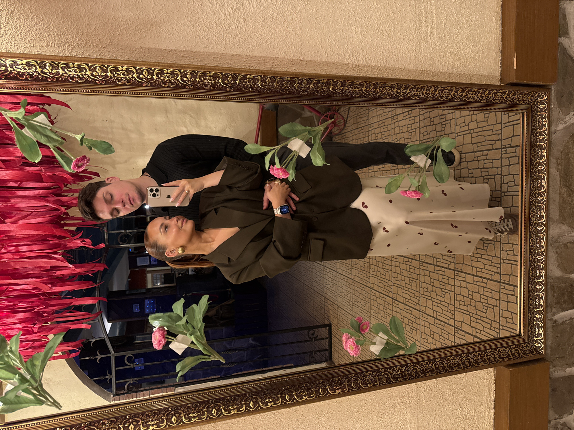 A reflection in a large, ornate, gold-framed mirror shows a couple taking a selfie. The man, wearing a black long-sleeved shirt and dark pants, holds an iPhone and looks at the screen. The woman, dressed in a dark green blazer and a long, light-colored dress with dark polka dots, looks away from the phone. The reflection behind them includes a brick-patterned wall, a red ribbon installation, and floral decorations attached to the mirror. The surrounding environment appears to be indoors with neutral-colored walls. The location in Chaikovsky, Russia cannot be confirmed from the image.