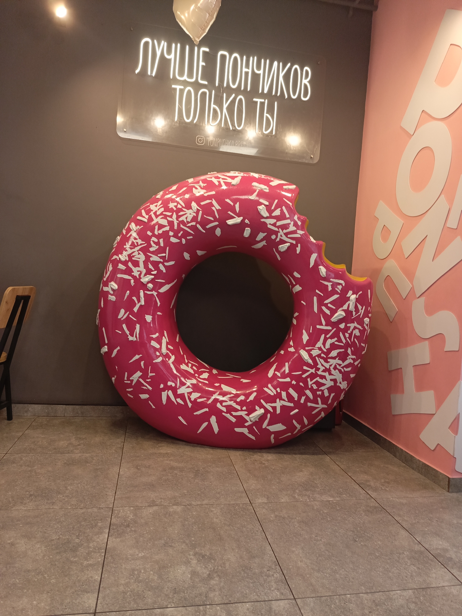 In Brest, Belarus, a large, pink, donut-shaped sculpture with white sprinkles is displayed indoors. Above the sculpture, a neon sign in Russian reads "ЛУЧШЕ ПОНЧИКОВ ТОЛЬКО ТЫ" (Only you are better than donuts). Below the neon sign, an Instagram logo and handle "@PONPUSHKA.BREST" are visible. The sculpture is positioned against a dark grey wall, with a wooden chair with a black metal frame to its left. The floor is tiled with large, square, grey tiles. To the right of the sculpture, a pink wall features large white cut-out letters that appear to spell out "DONUSHKA". The scene is illuminated by artificial lighting.