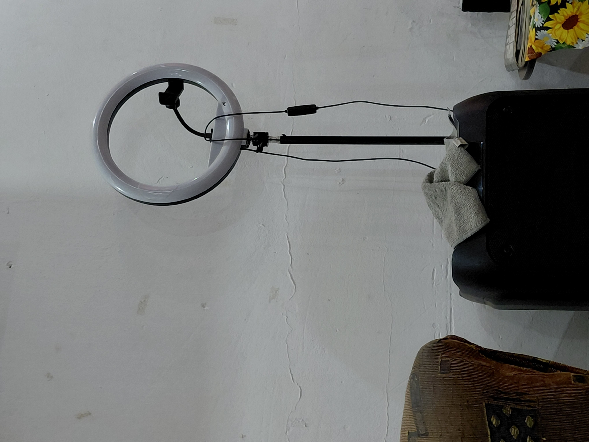 The image displays an indoor scene, predominantly featuring a white, textured wall against which various objects are arranged. The overall orientation suggests a vertical composition, possibly of a wall-mounted or freestanding setup.
On the left side of the frame, a prominent white ring light is visible, mounted on a black stand. Several black electrical cables extend from the ring light, running horizontally along the stand towards the right. One of these cables incorporates a small, black rectangular component, likely a controller or power adapter.
The ring light stand appears to be attached to, or resting upon, a large, black, rectangular electronic device, which resembles a speaker, positioned on the right side of the image. A light grey, textured cloth or towel is casually draped over the top right edge of this black device.
Above the electronic device, a small section of a fabric with a vibrant yellow sunflower and white daisy pattern on a dark background is visible. In the bottom right corner, a segment of a brown, intricately carved wooden object is seen. This object exhibits distinct geometric patterns and a coarse texture, suggesting it could be a piece of furniture, such as a stool, or a decorative drum, possibly reflecting cultural aesthetics.
The white wall serving as the background shows signs of age or wear, including minor vertical cracks and varied surface textures. There are no people visible in the image, and no specific activity is taking place; the scene depicts a static arrangement of items. The lighting appears to be ambient indoor light, making the precise time of day indeterminate. No visible text is present in the image. The presence of the ring light suggests a setup potentially used for photography, video recording, or online communication within a residence or workspace in Santa Ana, Venezuela.