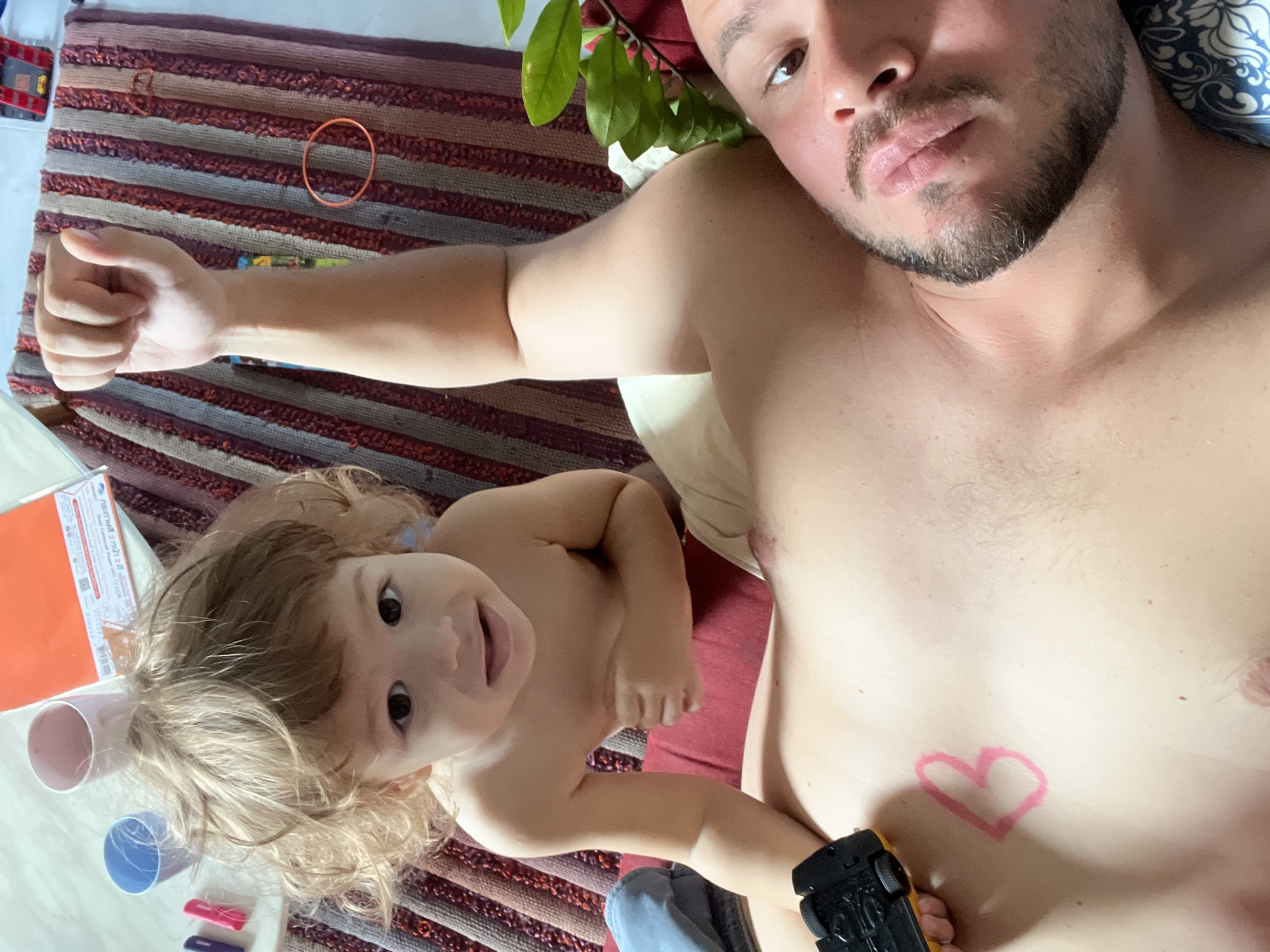 This media file shows a close-up, overhead view of an adult man lying down with a young child on his chest. The man is shirtless and has a pink heart drawn on his chest. The child has blonde hair and is looking up at the camera with a smile. The background is a striped rug.
The scene appears to be casual and intimate, likely taking place indoors. There are no strong indicators of time of day or weather. The emotion conveyed is one of affection and playfulness.
Objects visible include the striped rug, a pink cup, a blue cup, a yellow toy car held by the man, and what appears to be a notepad or book with an orange cover. There is also some greenery from a plant visible near the man's head.
No text is clearly legible in the image.