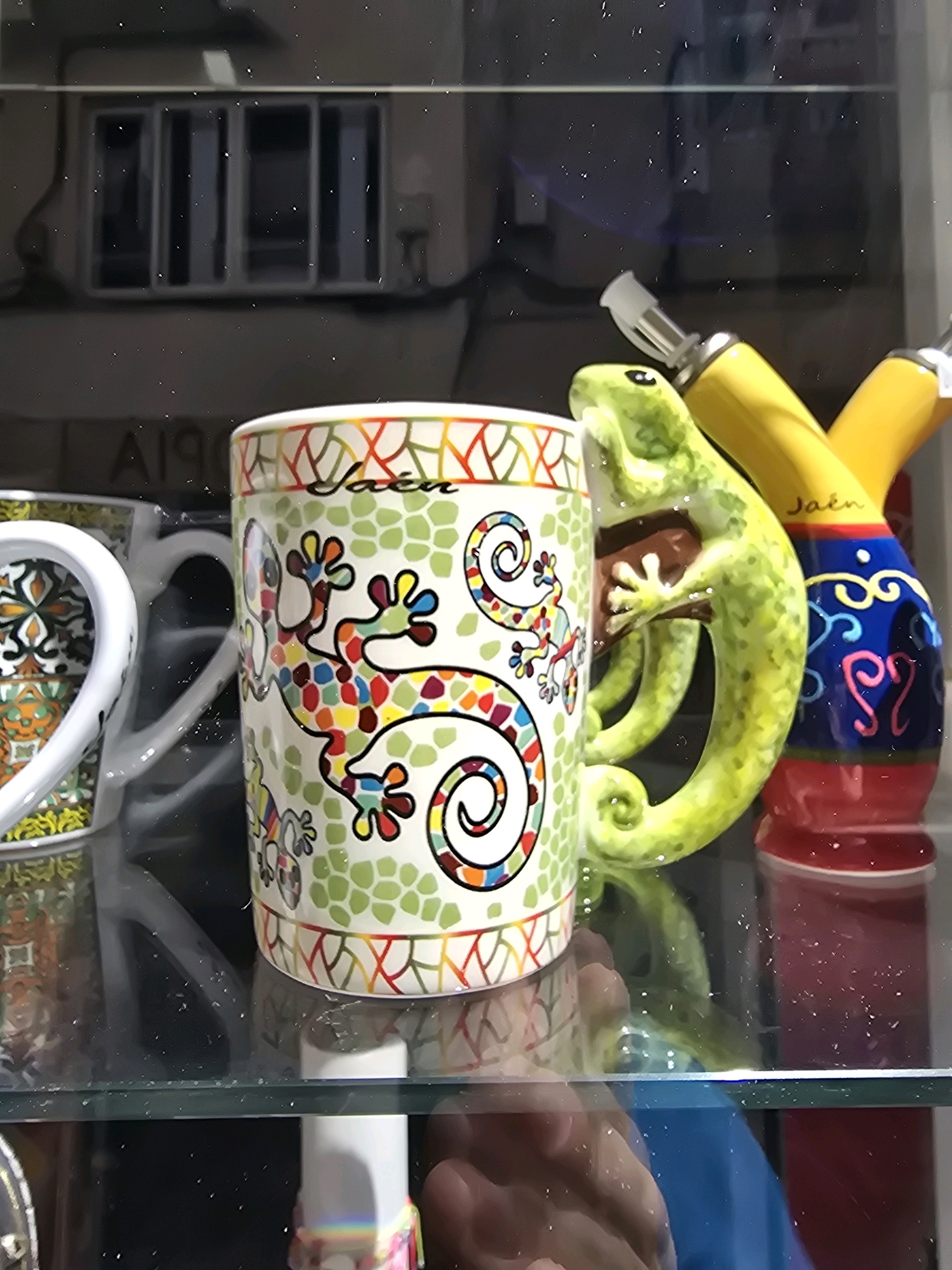 A close-up shot features a white ceramic mug decorated with a colorful mosaic pattern of lizards, set against a blurred background. To the right of the mug is a green ceramic lizard figurine, its body curving around the mug. Behind these items, a yellow object with a white tip and a red, blue, and yellow patterned cylindrical object are visible. The items appear to be displayed on a glass shelf, reflecting the surrounding environment. The word "Jaén" is visible on the mug, indicating its origin or association with the city of Jaén, Spain.