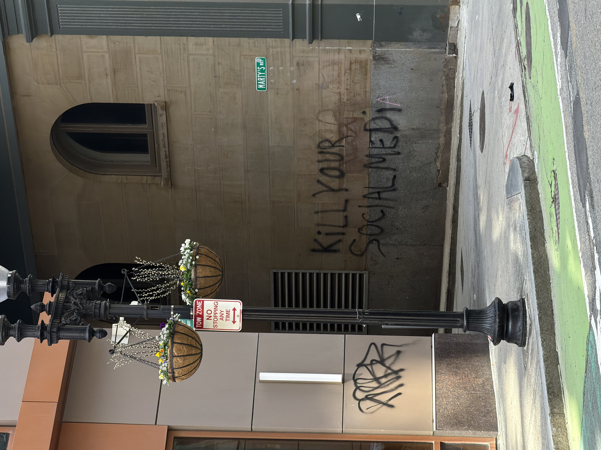 The image depicts a city scene, likely in Boston, United States of America, based on the "Marty's Way" street sign. The photo is taken at street level, with a lamppost in the foreground adorned with hanging flower baskets. A building stands in the background, covered with graffiti reading "Kill Your Social Media." There's a tow zone sign attached to the lamppost. Additional graffiti is seen on the lower section of the adjacent building. The street and sidewalk are visible, with green striping.