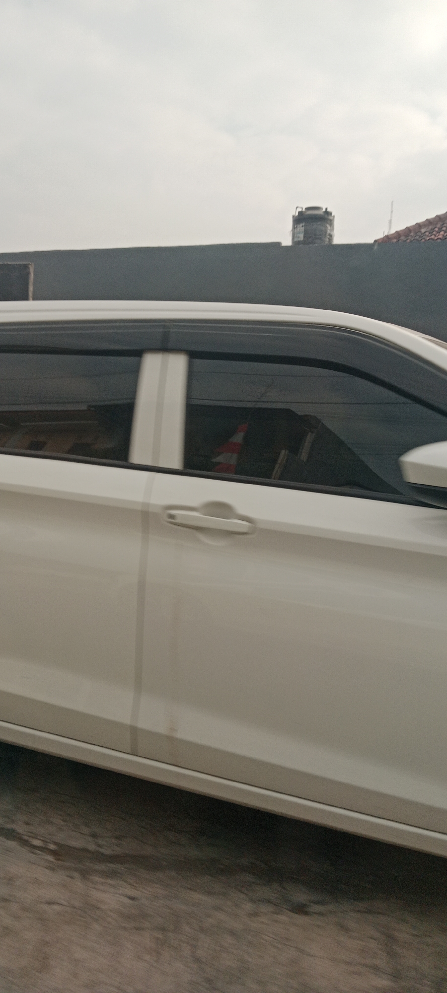 A white car is seen from the side, moving on a road. The car's windows are tinted, and there are vertical white stripes on the door. In the background, a gray wall is visible, with a water tank on top of it and part of a tiled roof. The sky is overcast. A traffic cone with orange and white stripes is reflected in the car's window. The scene appears to be in an urban environment in Maguwoharjo, Indonesia.