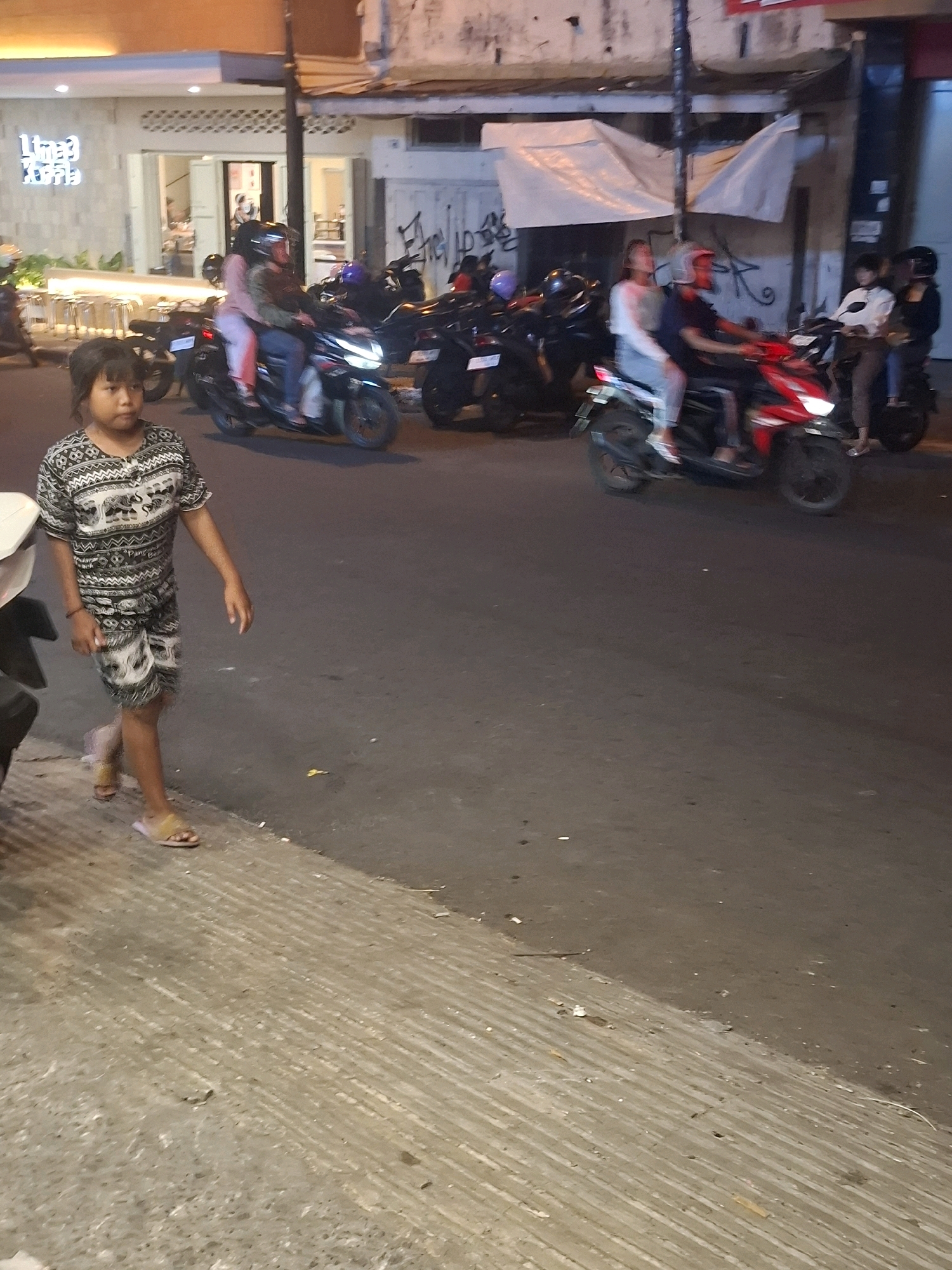 The image depicts a street scene in Cianjur, Indonesia, at night. A young girl in a patterned shirt and shorts walks on a sidewalk on the left side of the frame, with her left foot on the road. She is wearing sandals. Behind her, several motorcycles are parked. In the background, a building with an illuminated sign that reads "Limpa Coffee" is visible. Other motorcycles with riders are on the road, some of which appear to be moving. The street is paved and appears to be a commercial area.