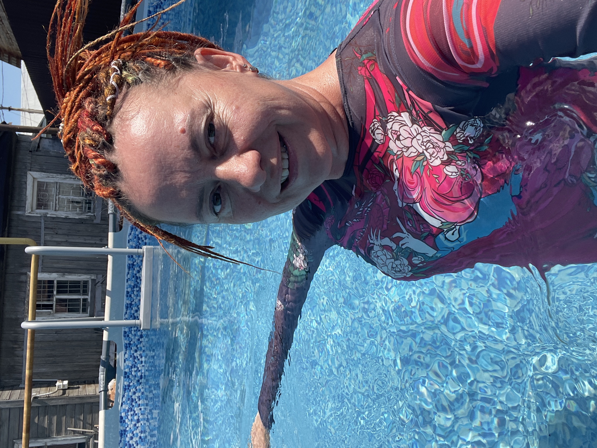 A woman with brightly colored synthetic dreadlocks is taking a selfie in a swimming pool. She is wearing a long-sleeved rash guard with a vibrant floral and dragon print. Her left arm is extended, and she appears to be holding onto the edge of the pool. The water is clear blue with sunlight creating shimmering patterns on the surface. In the background, there is a weathered wooden building and a portion of a swimming pool ladder. The scene suggests a casual, outdoor setting, likely during daytime. There is no information to indicate this location is in Orenburg, Russia.