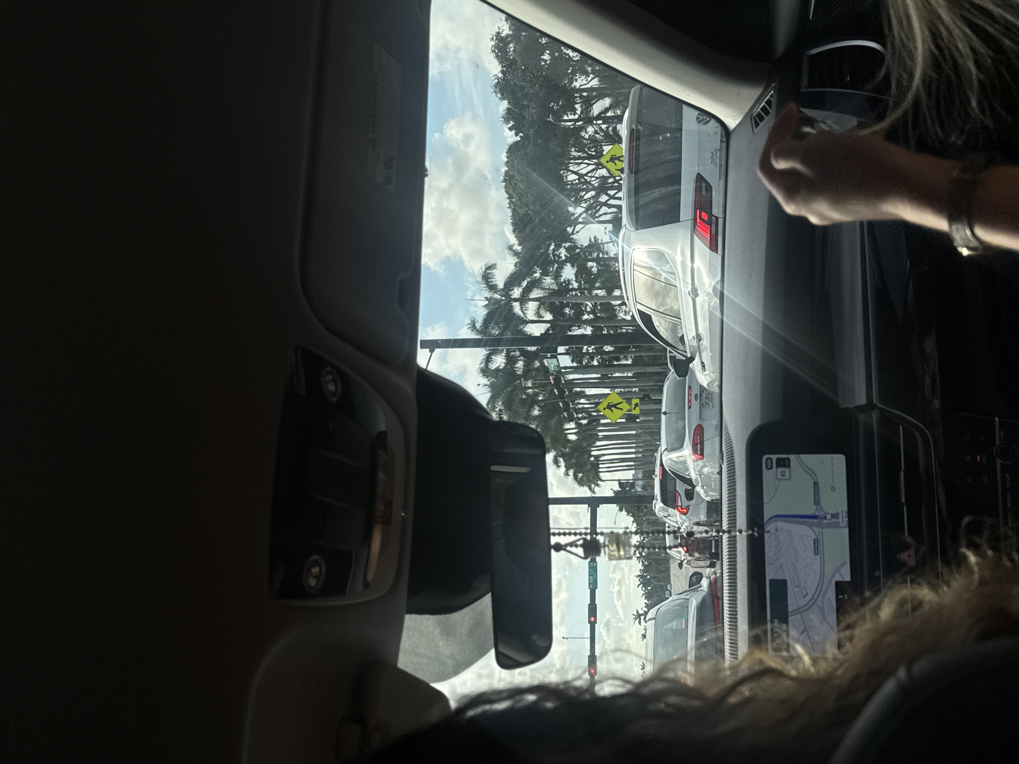The image, taken from inside a vehicle, depicts a view through the windshield and side window, revealing heavy traffic. Several white or light-colored cars are visible in front, with palm trees lining the road and clear skies in the background. Street signs, including a "pedestrian crossing" sign, are visible. The vehicle's navigation system displays a map. A person's hand is visible in the upper right corner, interacting with the vehicle's interior. The overall environment suggests a suburban area experiencing traffic congestion, likely in Boca Raton, based on the presence of palm trees and the navigation system display.