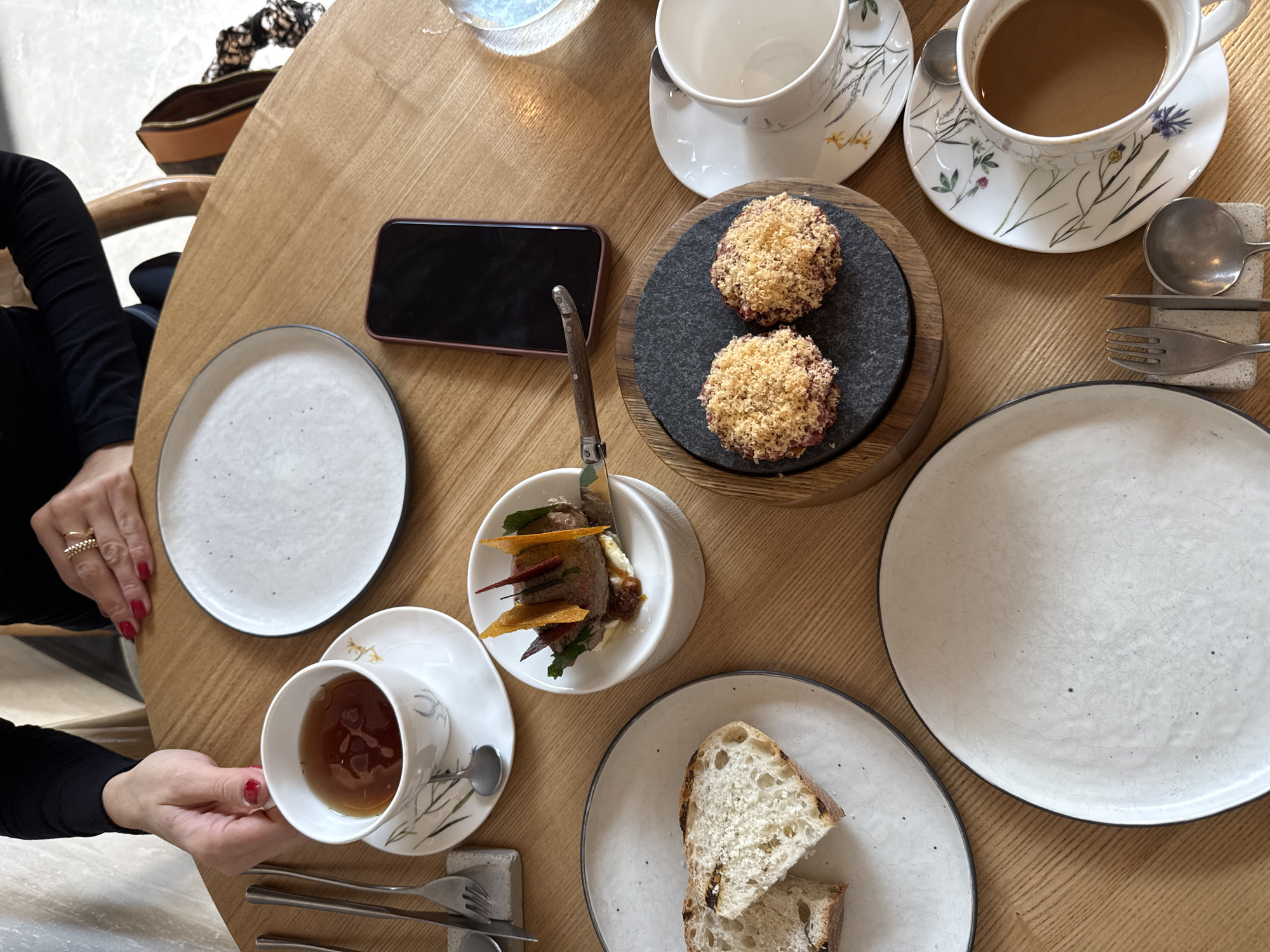 A high-angle shot depicts a wooden table at a restaurant setting. A woman, dressed in black with red nail polish and multiple gold rings on her fingers, holds a teacup filled with tea. The table features two empty plates, one plate with two slices of bread, and a dish with what appears to be beef tartare, topped with fried vegetable strips, garnished with herbs. Nearby is a black phone, two small, round, breaded meatball-like appetizers on a circular dark grey slate plate, and two floral teacups and saucers: one empty and one filled with coffee. Silverware is present on either side of the bottom plate. The scene is captured indoors with bright natural light, giving it a clean and modern aesthetic. There is no visible signage to confirm that this image was taken in Krasnoyarsk, Russia. Therefore, that information can not be verified from this image.