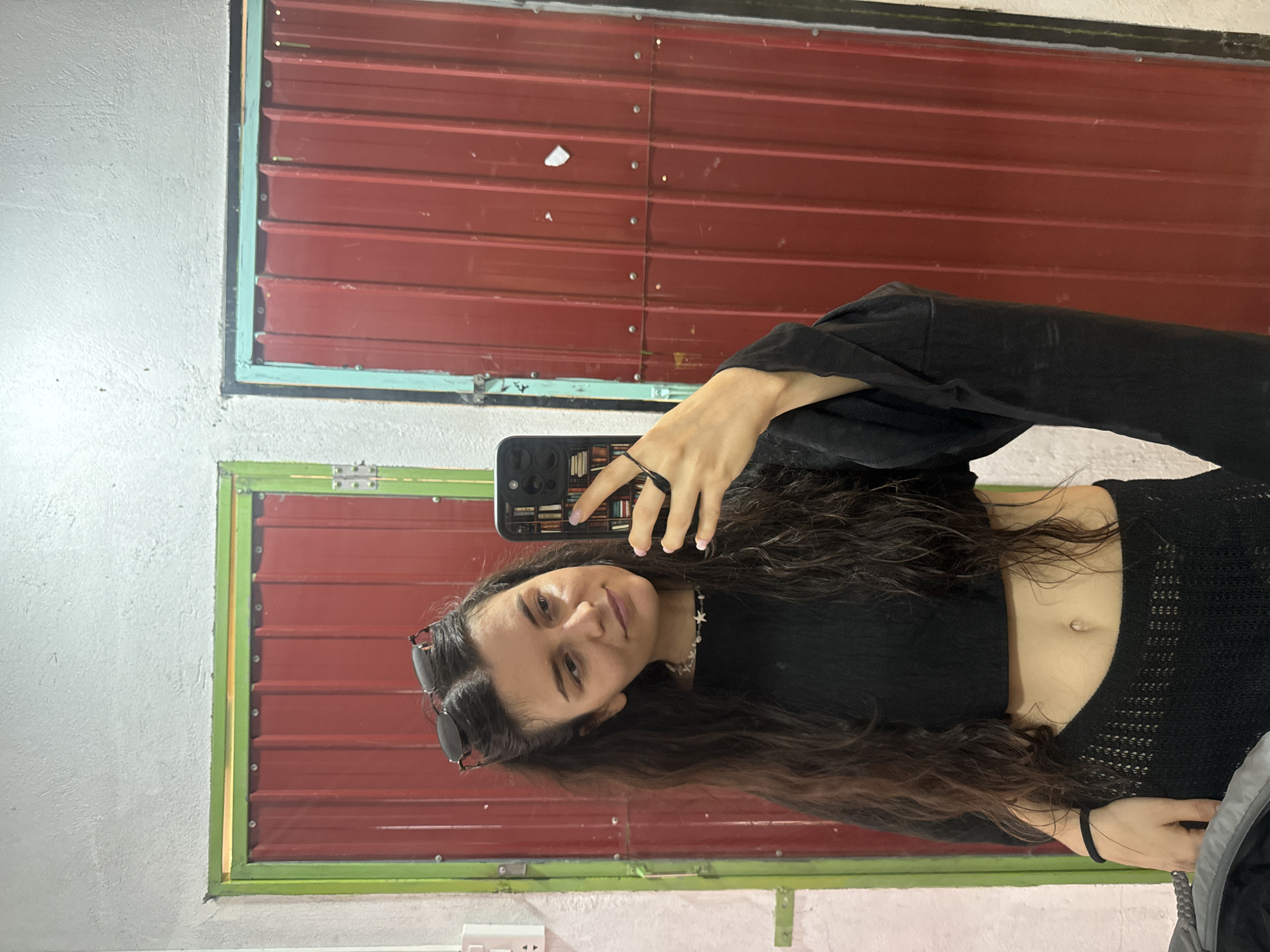 A young woman is posing for a selfie in front of a wall with two windows that have red metal louvered shades. The woman is wearing a black crop top and has long, dark wavy hair. She is holding a smartphone in her left hand and has sunglasses on her head. The wall appears to be made of concrete and is painted white. The windows have frames painted in a light green color. The location is likely in the city of Mũi Né, Vietnam, given the presence of similar architectural features.