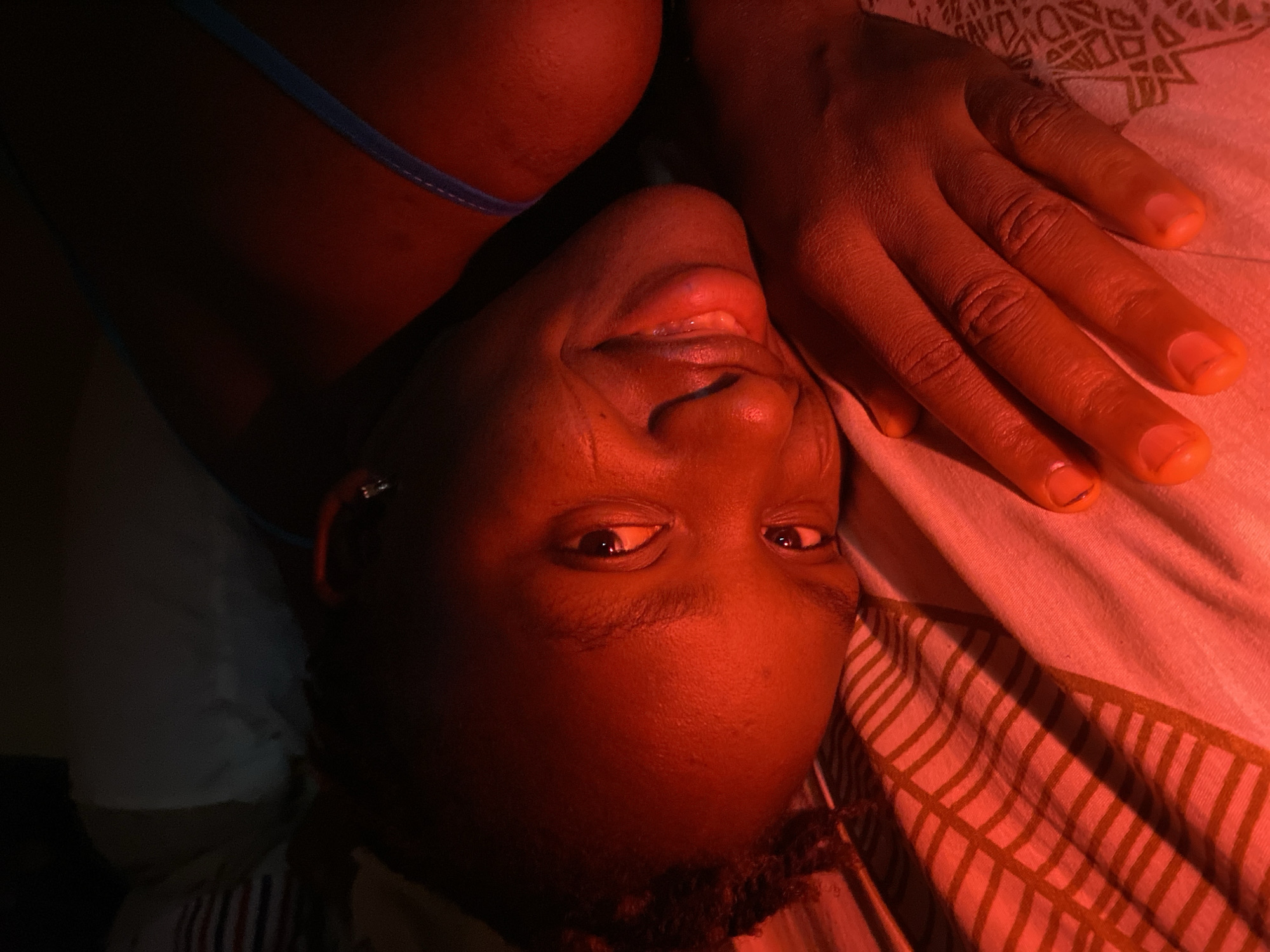 A person lies on a bed with striped bedding. They are illuminated by a red light source, and the location is unspecified, not linked to Kuru Dungus, Nigeria. The person's right hand rests on the bed beside their face. They are wearing what appears to be a blue strap, suggesting clothing. They have an earring in their left ear. They are looking at the camera and smiling with their mouth slightly open.