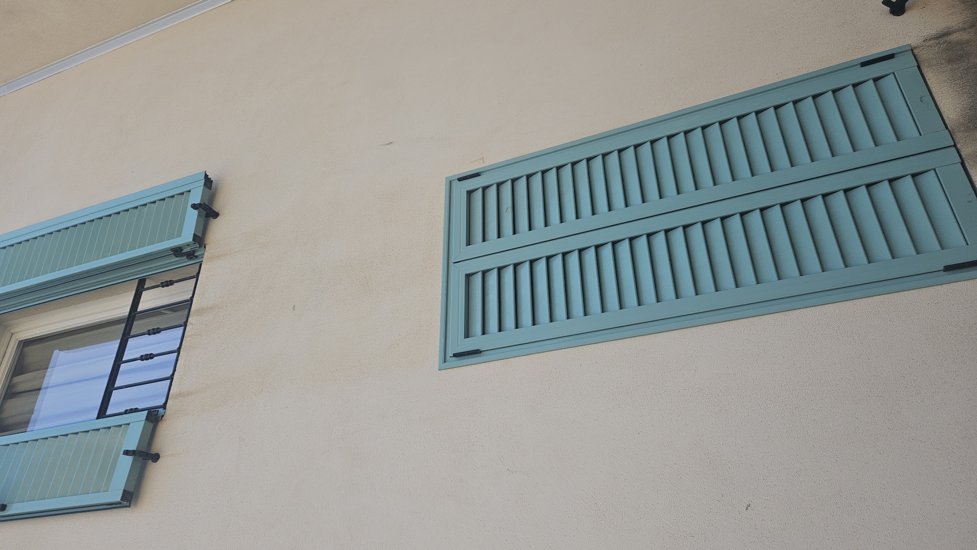 The image shows a building facade in L'Île-Rousse, France, with two windows fitted with teal-colored louvered shutters. The building's wall is a light beige, textured stucco. The upper window is partially open, revealing a glimpse of a curtain inside and a black metal railing below it. The lower window is entirely covered by the closed teal shutters. The perspective is from below, looking up at the building, with the shutters angled relative to the camera. The lighting suggests it is daytime, with shadows cast on the wall. There are no people or animals visible in the image.