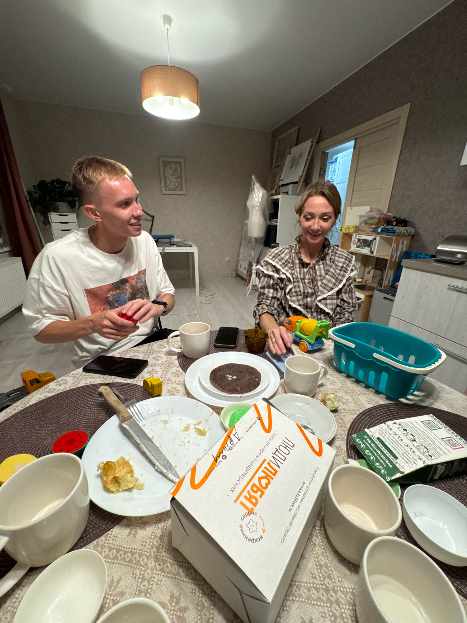 The image shows a dining table in what appears to be a residential setting, possibly in Saint Petersburg, Russia. A young man with blonde hair and a white t-shirt is seated at the table, holding a small red object and looking to his right. Across from him, a woman with brown hair, wearing a checkered blouse, is also seated and looking towards the man. The table is covered with a patterned tablecloth and is laden with used dishes, cups, plates with remnants of food, and several small toy vehicles. A teal plastic basket is also on the table. In the background, there is a kitchen counter with appliances, shelves with various items, and a doorway. The lighting suggests an indoor environment, likely during the evening.