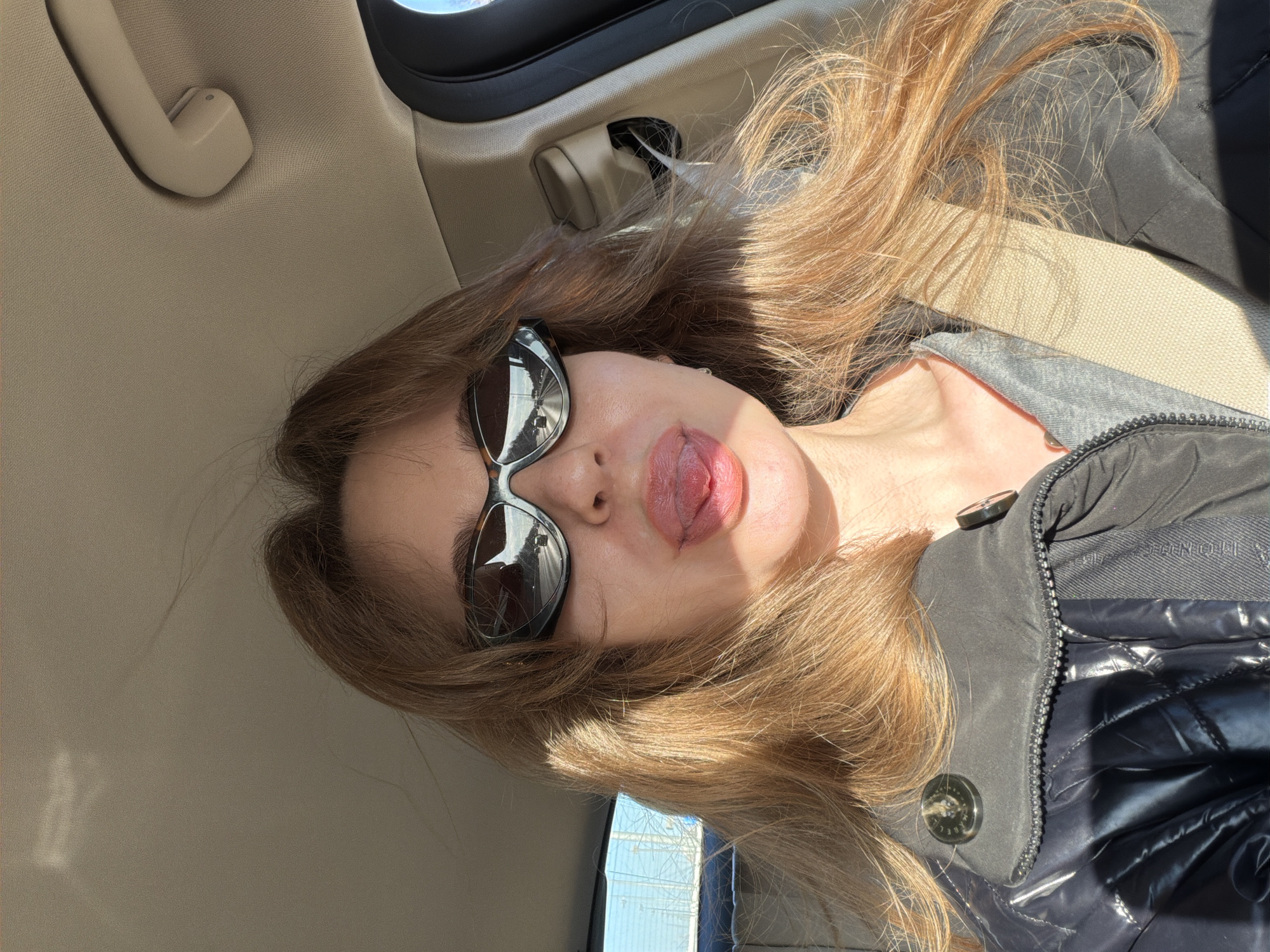 A woman is depicted in a close-up selfie, likely taken inside a vehicle. She is wearing sunglasses and has her tongue slightly protruding. The interior of the car is visible, including the ceiling and a grab handle. The woman is wearing a dark-colored puffer jacket. It is not possible to determine her exact location as Moscow, Russia from the image.