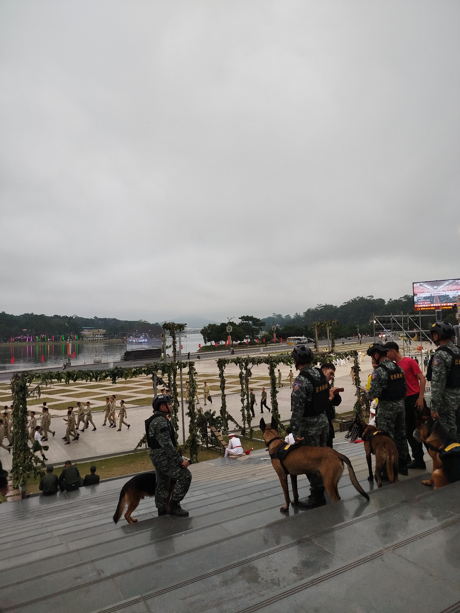 The image depicts an outdoor scene in Da Lat, Vietnam, likely near Xuan Huong Lake, a popular tourist destination. The foreground features a set of wide, dark gray stone steps. Several individuals in military-style uniforms are positioned on and around these steps. Some of these individuals are handlers with four dogs. The dogs appear to be Belgian Malinois and German Shepherds, some wearing protective gear like muzzles and vests with the marking "CSDN" (likely Canine Security and Detection Unit).
In the mid-ground, a paved area is visible, with more people in uniform, possibly cadets or participants in a ceremony, marching or walking in formation. Beyond this area, Xuan Huong Lake stretches across the frame, its surface reflecting colorful lights from decorations on the shoreline. Trees line the far bank of the lake, and in the distance, rolling hills and mountains can be seen under a cloudy sky. A large screen displaying images and text is visible on the right side of the frame, suggesting an event or performance is taking place. The overall atmosphere is formal and organized, possibly a security detail for a public event.