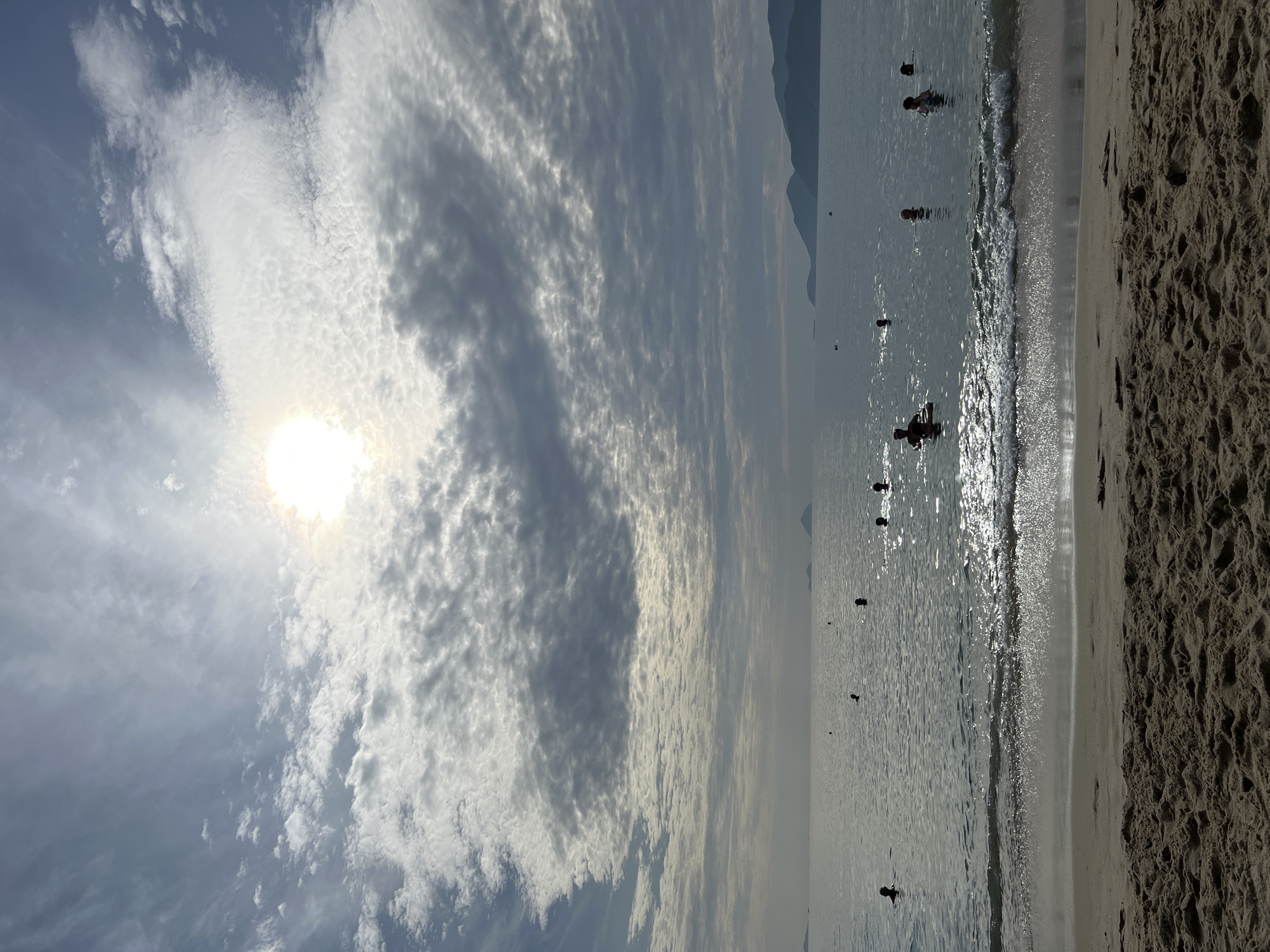 The image depicts a serene beach scene at Nha Trang, Vietnam. The sun, partially obscured by a dramatic, textured cloud formation, casts a bright glow over the calm sea. The water is populated by several individuals swimming or wading, their dark silhouettes contrasting with the sunlit surface. In the foreground, the sandy beach shows the subtle textures of dried sand. Distant hills and islands form a gentle silhouette against the hazy horizon. The overall atmosphere is one of peaceful recreation on a slightly overcast day.