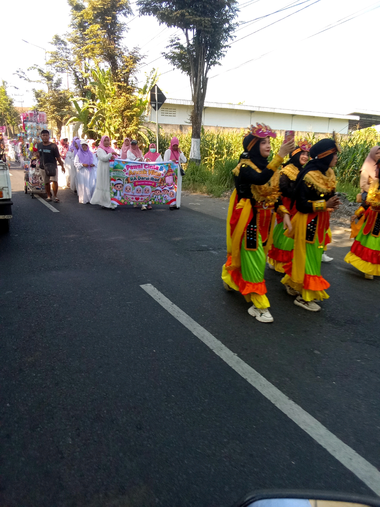 In Pancakarya, Indonesia, on a sunny day, a group of people are participating in a parade on a paved road lined with trees and buildings. Several individuals are wearing traditional attire, including women in white hijabs and colorful dresses, and performers in vibrant yellow, green, and orange costumes with elaborate headwear. Some are holding a banner that reads "Pawai Cinta & Anca Hias RA Darul Ibad". A man in a dark t-shirt and shorts is walking alongside a cart, and others are visible in the background. The road has a white line marking its edge. The overall scene suggests a cultural or festive event.