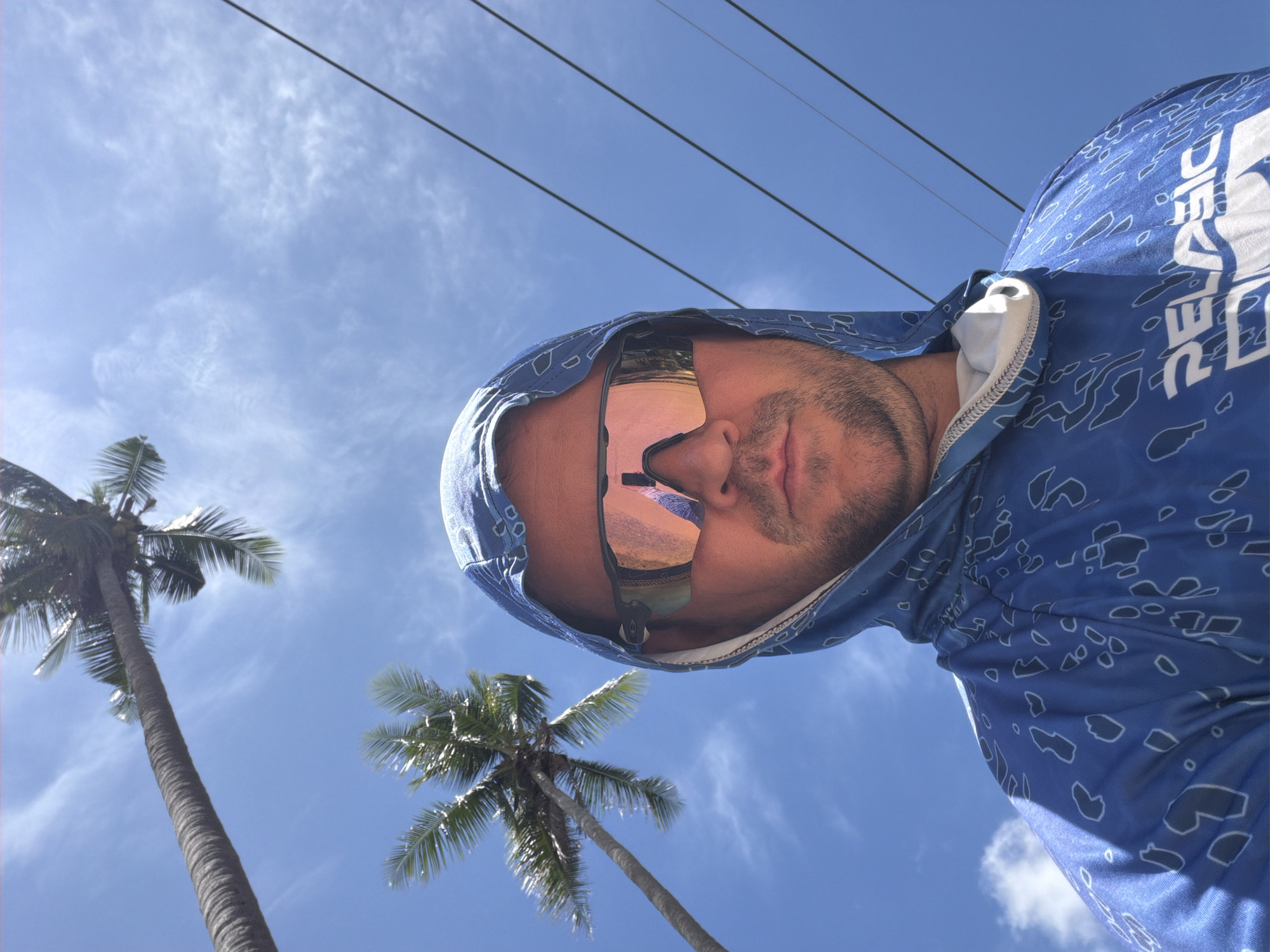 A man wearing a blue hooded shirt with a white pattern and mirrored sunglasses poses for a selfie outdoors under a bright blue sky. He has short brown hair and a beard. Two palm trees are visible in the background, with their fronds reaching towards the sky. Power lines are also visible against the sky. The man is wearing a blue shirt with a white graphic logo that reads "PELAGIC". The image appears to have been taken in Baan Bo Phut, Thailand.
