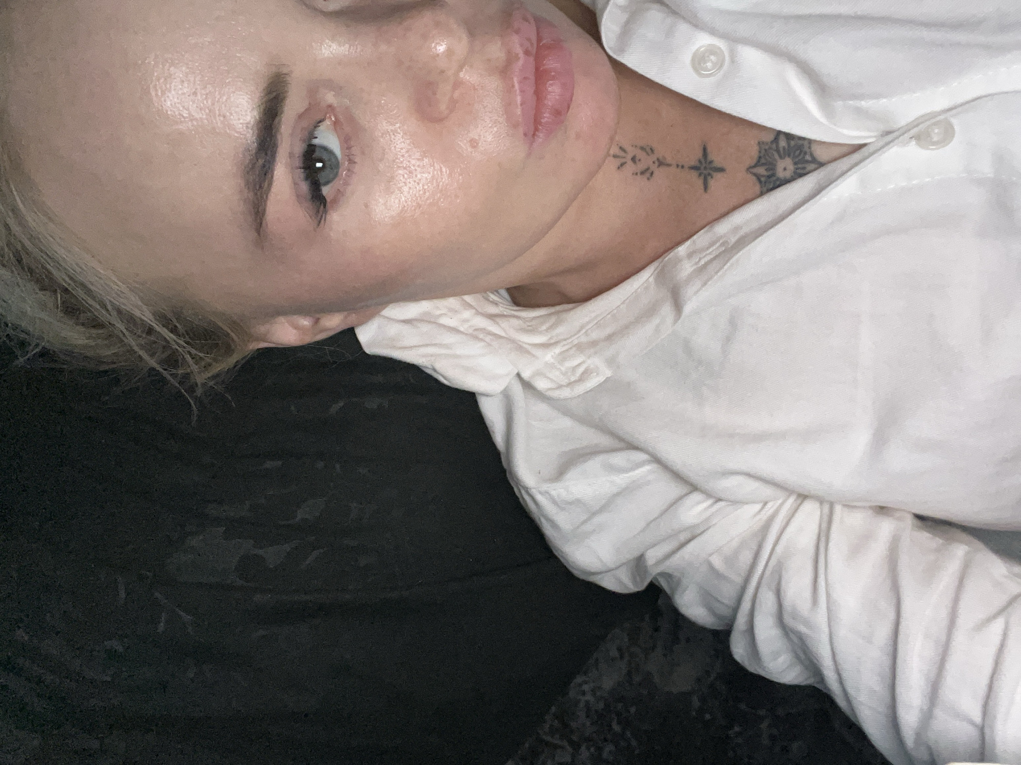 A close-up, waist-up shot shows a fair-skinned woman with blonde hair pulled back, wearing a white button-down shirt. She has light blue eyes, defined eyebrows, and is wearing light makeup. Her lips are slightly parted, revealing what appears to be lip filler. She has two tattoos on her neck: a star-like design above a circular, mandala-like tattoo. The background is dark and out of focus, with textures suggesting fabric or a dark room. The lighting is somewhat dim, highlighting the sheen on her skin.