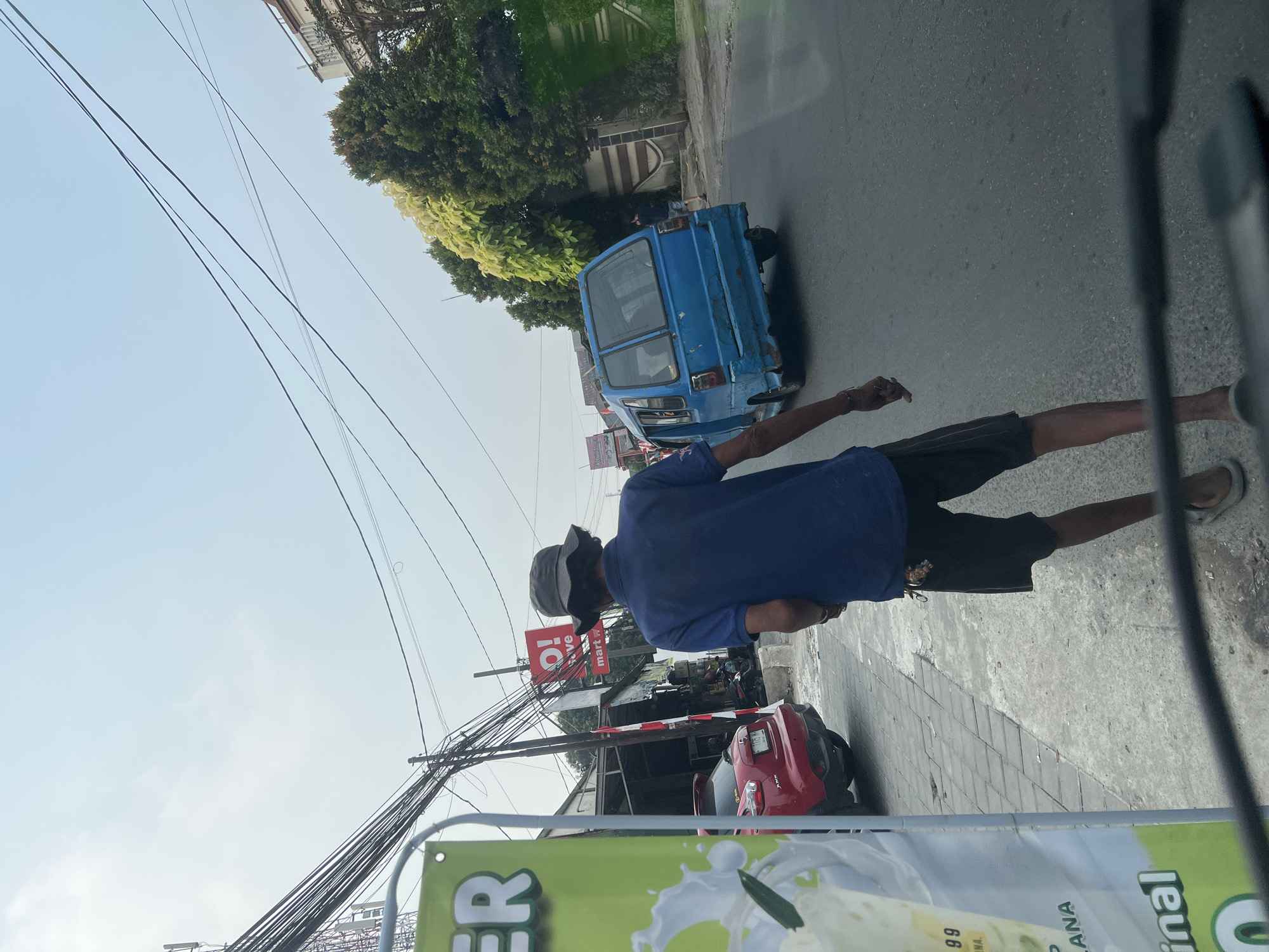 The image depicts a street scene in Citeureup, Indonesia. In the foreground, a person wearing a blue t-shirt, dark shorts, and a bucket hat is walking away from the camera on a sidewalk. The person's left arm is extended slightly, and they are wearing sandals. To the left of the person, a large banner with green and white lettering is visible. Behind the person, a blue minivan is driving on the road. Further down the street, a red car is parked on the side. The environment appears to be an urban or semi-urban area with buildings, trees, telephone poles, and overhead wires. The sky is clear and bright.