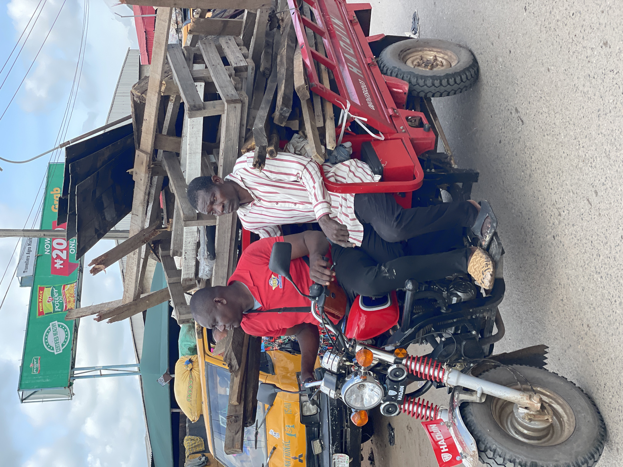 Two adult males are observed riding a red motorized three-wheeler, branded "HADJIN," on a paved road under an overcast sky. The front of the vehicle features a single wheel with a silver fender and red shock absorbers. The rear has two wheels supporting a red cargo bed heavily loaded with assorted wood pieces, including planks and broken pallets. A phone number, 08060508033, is partially visible on the cargo bed. The driver, wearing a red polo shirt, dark trousers, and textured brown slip-on shoes, holds the handlebars. The passenger, seated to the driver's right, wears a red and white striped long-sleeved shirt and dark trousers with dark sandals. In the background, a yellow van or bus, marked "NATIONAL" and "MITSUBISHI" with a license plate "LAGOS GZ62H," is visible behind the three-wheeler, with luggage on its roof. Above, a large green billboard advertises "Power Oil" and "CERTIFIED HEALTHY," with "Travellers Agro Mark" at the top. Adjacent to it, another billboard displays "NOW #20 ONLY" and "Grab," indicating a commercial area in Lagos, Nigeria.