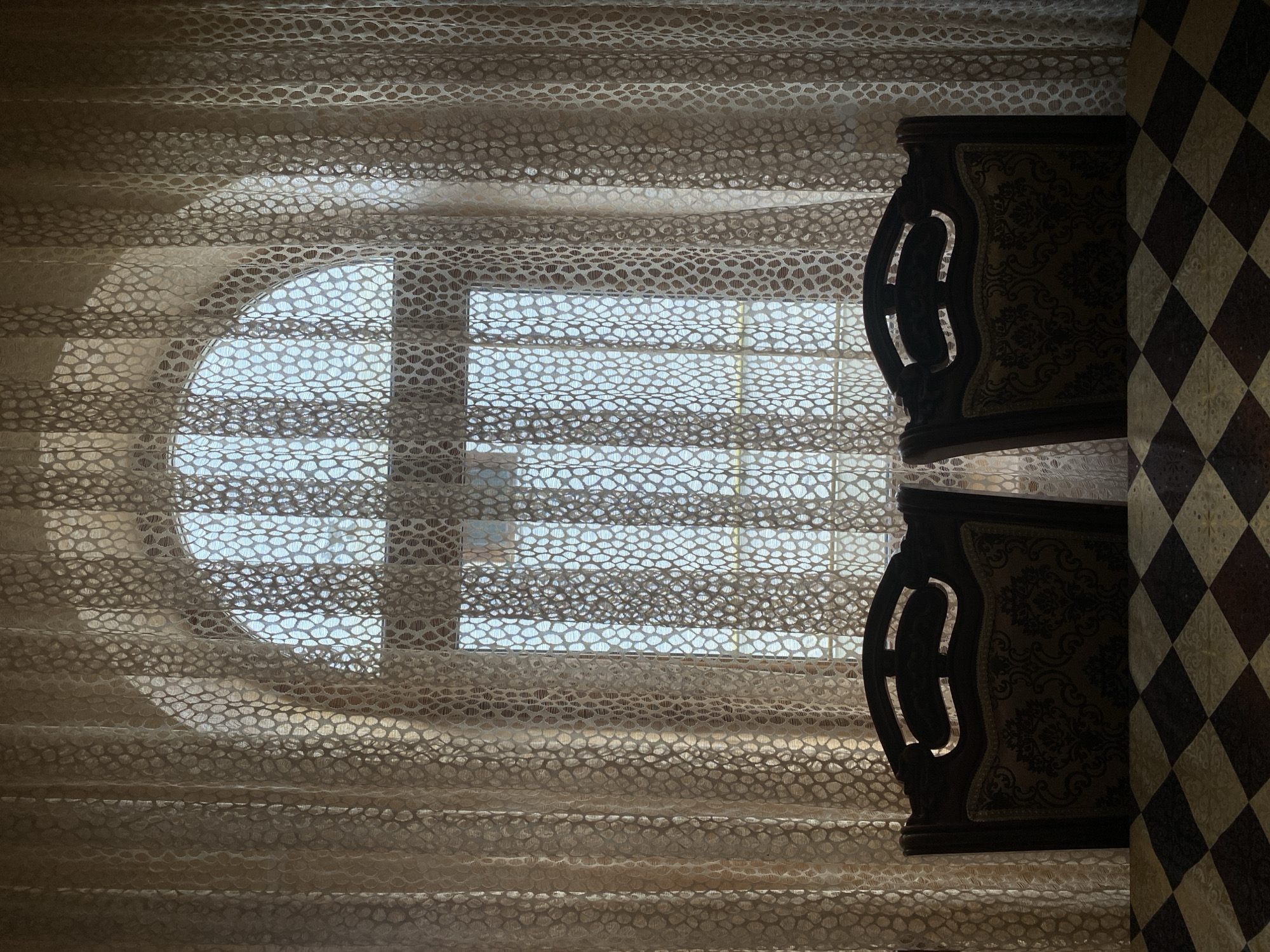 The image displays an interior view of a room, likely a dining area, looking towards an arched window in the city of Unknown, Unknown. The window is predominantly obscured by two panels of light beige, sheer curtains featuring a repeating, organic, pebble-like pattern. Bright daylight from outside illuminates the curtains, revealing a light blue sky or distant water through the translucent fabric. In the mid-ground, two dark wooden dining chairs, each with a patterned upholstered backrest and a carved top rail with decorative cut-outs, are positioned with their backs to the viewer, facing the window. In the immediate foreground, the edge of a table is visible, covered by a tablecloth patterned with alternating dark brown and light beige squares. The interior is partially shadowed, creating a contrast with the brightly lit window. No individuals or active subjects are present.