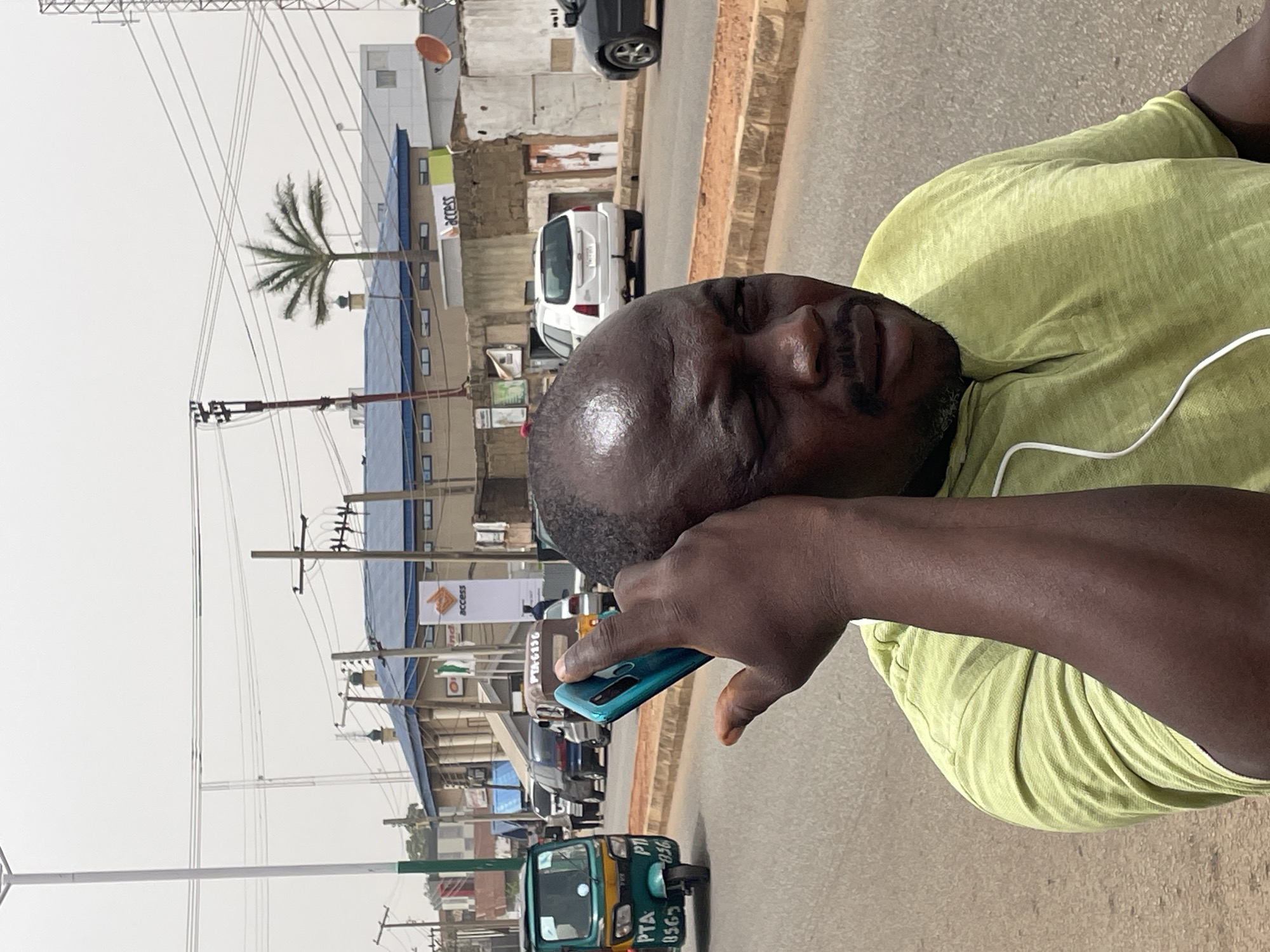 A close-up shot of a man with dark skin and a bald head, wearing a light green shirt and holding a turquoise smartphone, with his face and upper body taking up a large portion of the frame. He appears to be outdoors in the city of Jos, Nigeria. Behind him, there is a street scene with parked cars, a three-wheeled green auto-rickshaw, electrical poles and wires, and buildings, including one with an "Access" sign. A palm tree stands tall behind one of the buildings.