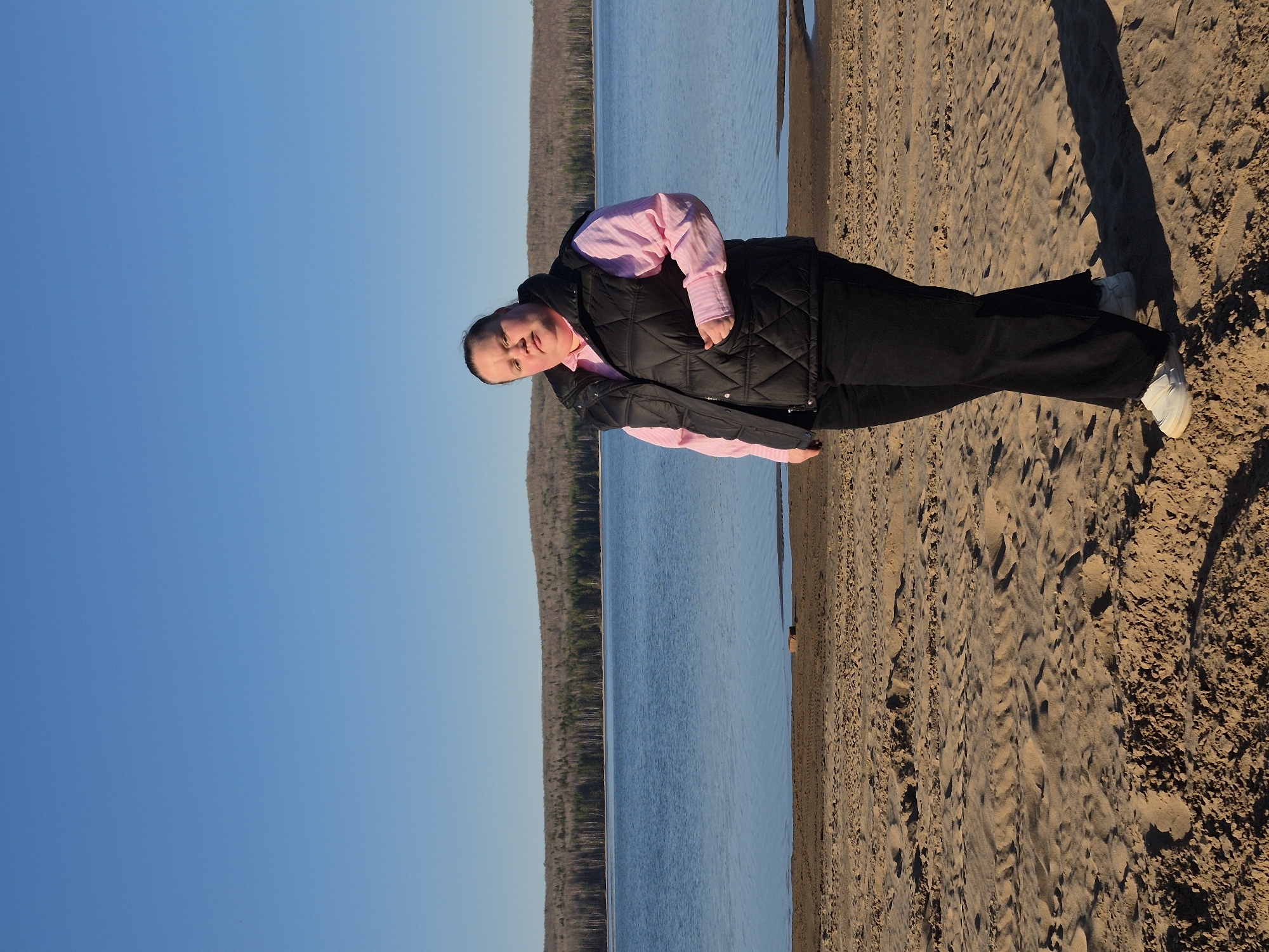 A person stands on a sandy beach next to a large body of water, possibly the Kama Reservoir, near Chaikovsky, Russia. The person is wearing a pink collared shirt, a black puffy vest, black pants, and white sneakers. They have their hands in their pockets. The sky is clear and blue. A forest line is visible across the water. The sand is textured with footprints and shadows, indicating a sunny day.