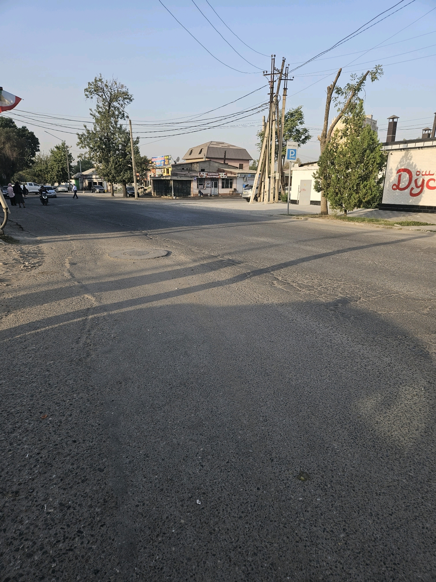 The image shows a street scene in Dushanbe, Tajikistan, likely in the vicinity of the "Duy" shopping center. The perspective is from the street looking towards a row of buildings and overhead power lines. The road surface is asphalt, with visible wear and tear, including patches and cracks. A circular manhole cover is present in the foreground, and shadows of trees and power lines are cast across the asphalt.
On the right side of the street, a white brick building with a large red logo spelling "Duy" is visible. This building appears to be a commercial establishment. To its left, a coniferous tree stands, and further left, a large tree with trimmed branches is prominent. Behind these trees, more structures are visible, including buildings with pitched roofs and some greenery. A white utility box with a red warning symbol is mounted on a pole near the trees. A blue parking sign with a white "P" is also present.
On the left side of the street, several vehicles are parked or driving. These include a white SUV, a motorcycle with a rider, and other cars. People are seen walking on the sidewalk. A tall tree with dense foliage is situated further down the street on the left. The sky is clear and blue, indicating daytime. The overall impression is a typical urban street in Dushanbe on a clear day.