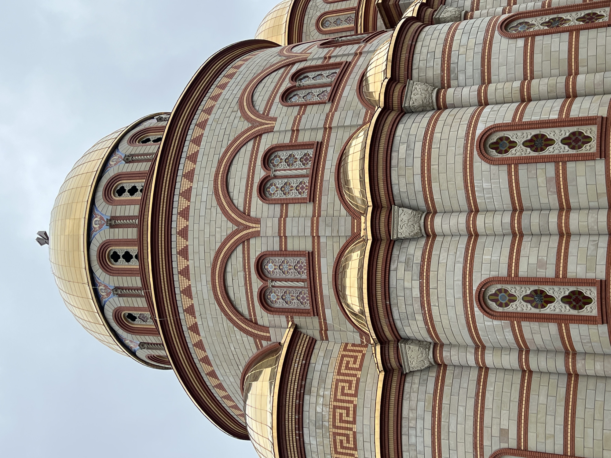 The image showcases the intricate architecture of the Cathedral of the Immaculate Conception of the Blessed Virgin Mary in Ternopil, Ukraine. The view is a low-angle shot emphasizing the building's height and detailed design.
The cathedral's structure features a combination of light-colored brick and red brick accents arranged in vertical stripes and decorative patterns. Notable elements include rows of arched windows with ornate floral-patterned designs, and golden domes that gleam against the sky. The domes are clad in gold-colored panels.
The sky is a uniform light gray, providing a neutral backdrop that highlights the architectural details of the cathedral.