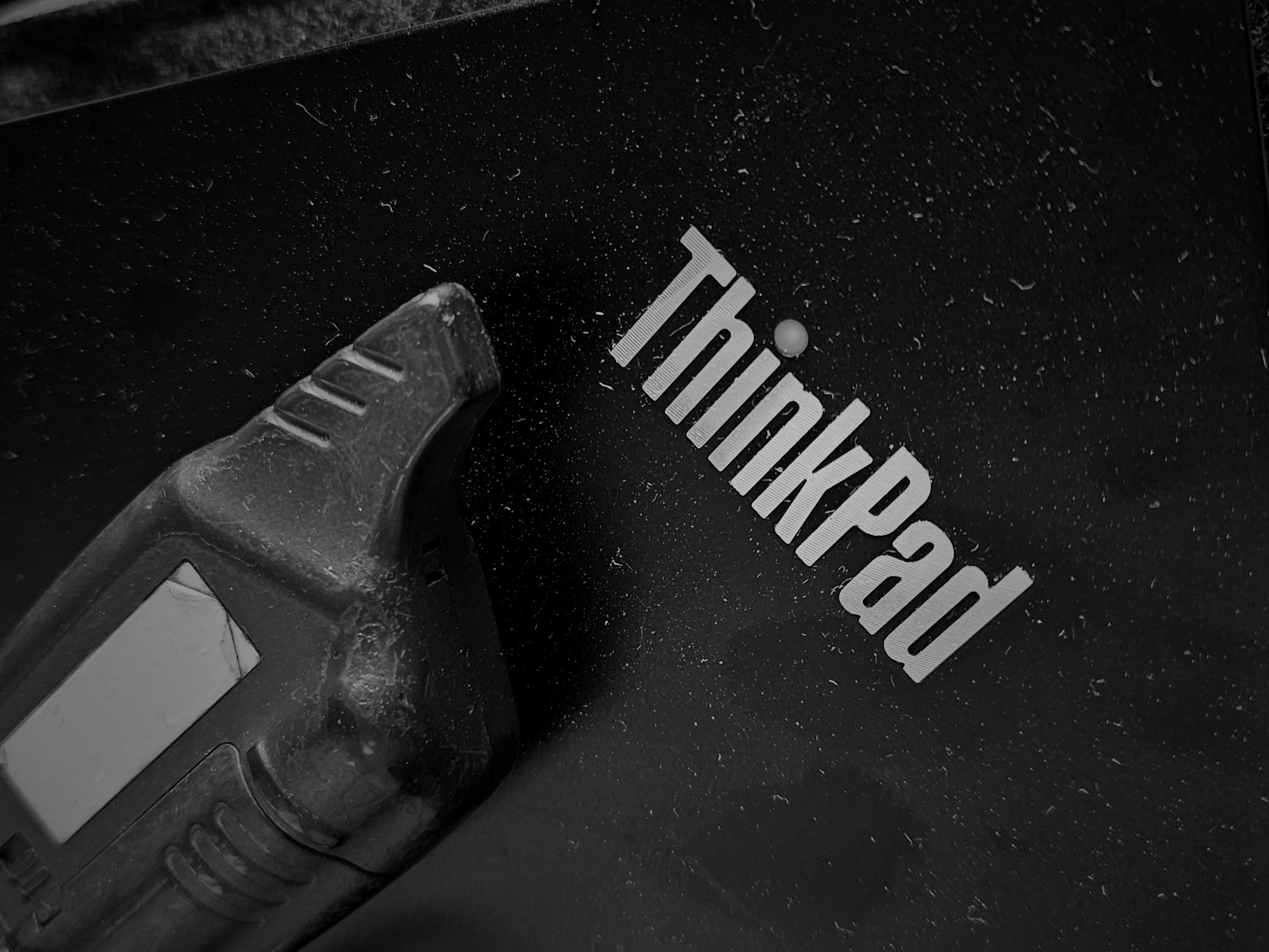 A black and white image shows a black ThinkPad laptop, partially covered with dust, with the "ThinkPad" logo visible in the upper right corner. To the left of the logo, partially obscuring it, is an ambiguous electronic device with a small display screen. The image exhibits a high degree of texture and visible detritus, suggesting the laptop and device have been exposed to environmental elements. The scene's location is not discernable from the provided image.