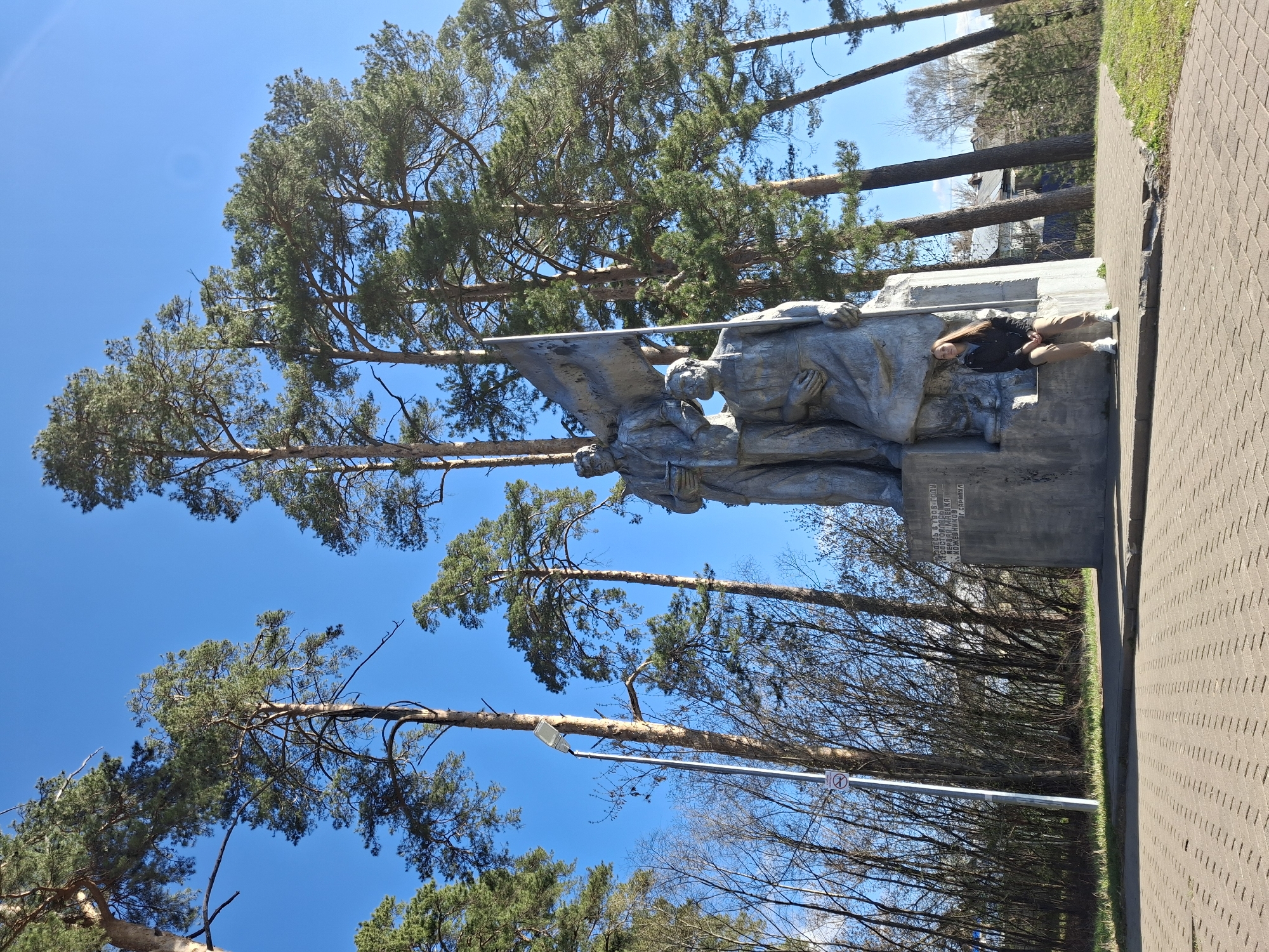 The image shows a war memorial in Sarapul, Russia. It consists of a stone monument featuring sculptures of two soldiers holding a flag. A young woman with long brown hair is lying on the memorial next to one of the soldiers. The memorial is located beside a paved walkway, and tall pine trees are visible in the background under a clear blue sky. A street light is also seen near the monument.