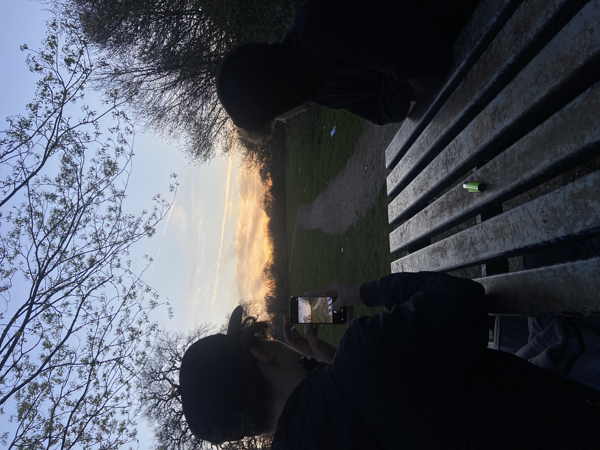Two people are sitting on a weathered wooden bench in a park at dusk or dawn. One person, wearing a dark hooded sweatshirt and a baseball cap, is holding a smartphone and taking a photo of the sunset or sunrise visible through the trees. In the background, a path leads into the distance through a grassy area. The setting is likely in Broxbourne, United Kingdom, given the prompt. A small, green object sits on the bench. The other person, wearing a dark hooded sweatshirt, is sitting next to the photographer. Bare branches of trees are visible against the sky.