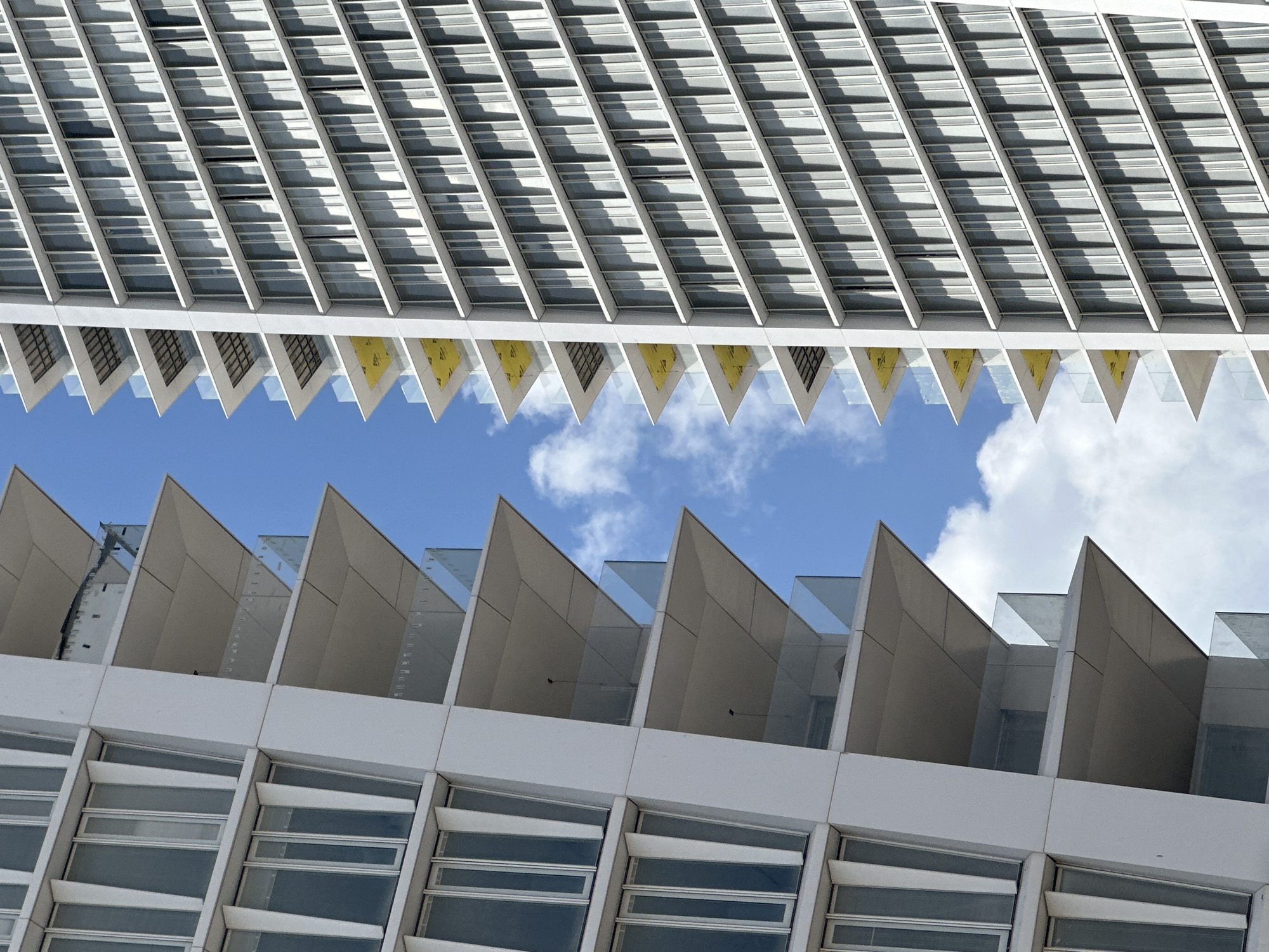 This close-up architectural photograph captures a modern building in Tel Aviv, Israel, seen from an upward perspective. The image is dominated by repeating geometric patterns and strong lines, framing a section of clear blue sky with scattered white cumulus clouds.
The upper portion of the frame features an inverted structure composed of numerous white and grey horizontal elements, possibly sun shades or louvers, which cast distinct diagonal shadows across their surfaces. Below these, a series of sharply pointed triangular forms creates a serrated edge. Some of these triangular elements reveal inner surfaces colored bright yellow.
The lower half of the image showcases another section of the building, featuring an upright series of similar angular, pointed structures with glass panels, forming a contrasting serrated profile against the sky. Beneath these, a facade with multiple rectangular windows is visible. These windows, set in white frames, appear to have horizontal louvered blinds or adjustable panels.
The bright lighting and sharp shadows indicate a sunny daytime environment with fair weather. The photograph highlights the abstract beauty of modern architecture, focusing on repetitive forms, light, and shadow. No people or activities are depicted, and no visible text is present.