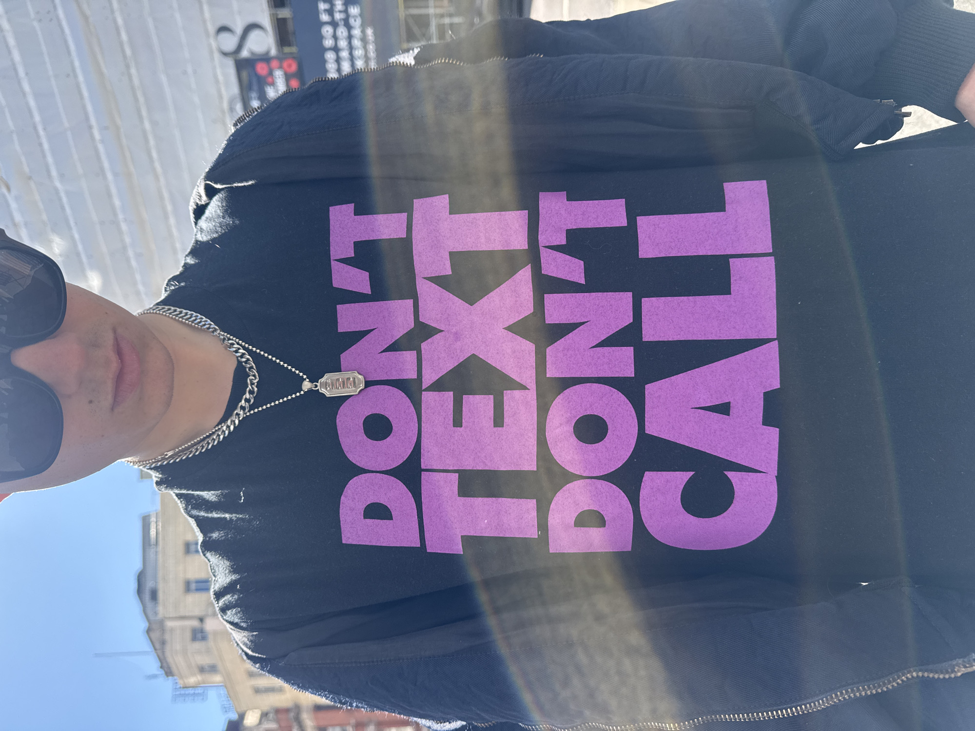 A person is depicted from the chest up in a close-up, upward-angled outdoor shot. They are wearing dark sunglasses, a black jacket, and a black t-shirt featuring prominent purple block letters that spell out "DON'T TEXT DON'T CALL." A silver chain necklace with a small, rectangular pendant, which has indistinct text resembling "RMMNL" or "RMWNL", is visible around their neck.
The image captures a bright, sunny day, indicated by the strong sunlight and a noticeable lens flare across the frame. The background reveals urban architecture, including light-colored buildings and a clear blue sky. Partially visible in the upper background is a commercial sign with fragmented text like "S", "SQ FT", "WARD-TH", and "KSPACE", suggesting a business or office space. The overall setting is a city environment, specifically London, United Kingdom. The person's expression is not visible due to the sunglasses, and the scene appears to be a casual moment captured in a photo.